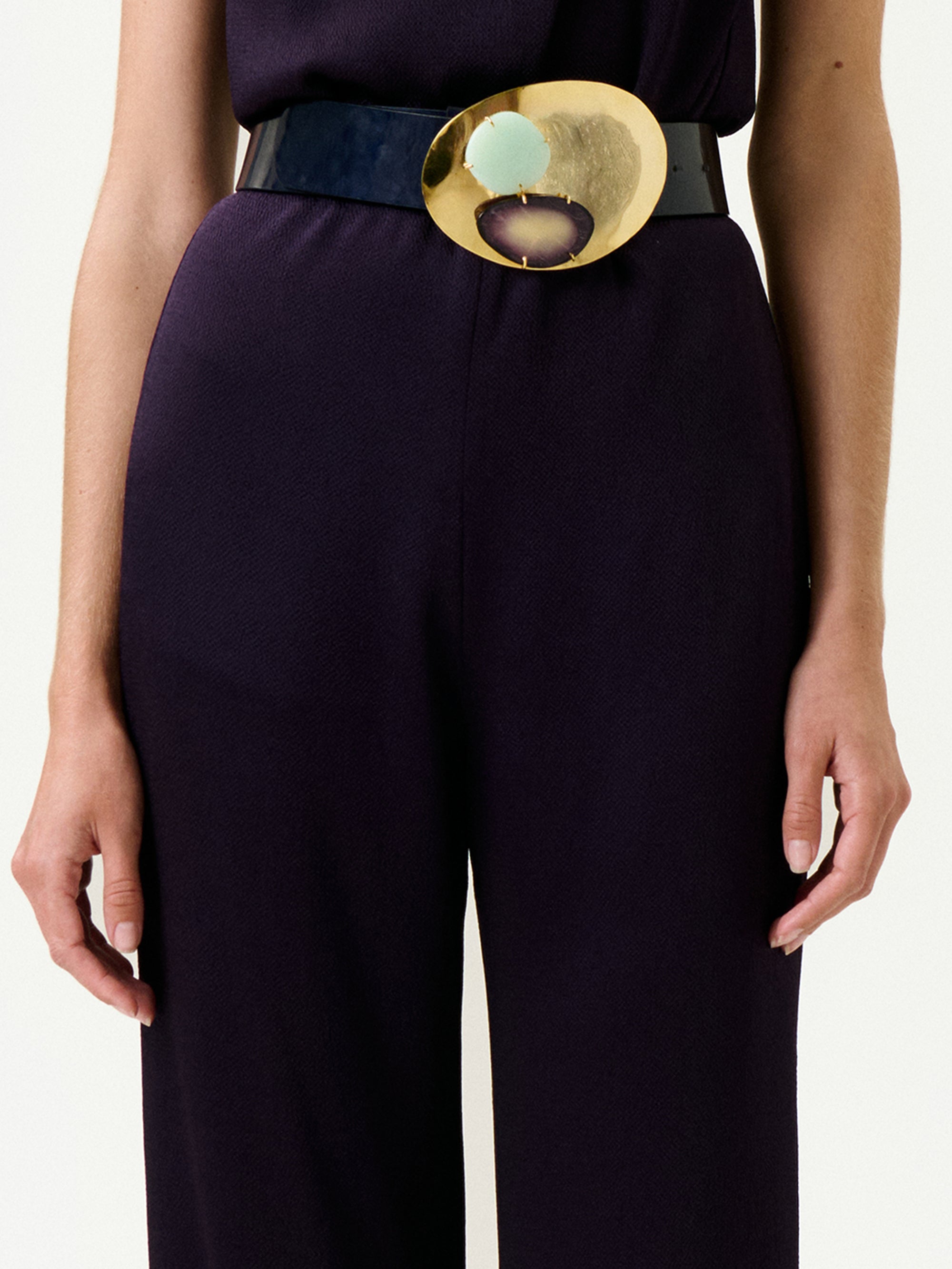 A person wearing the Palermo Pant in dark purple, styled with a true-to-size fit and a wide black belt featuring a large gold oval buckle with green and purple stones. Their arms are relaxed by their sides.