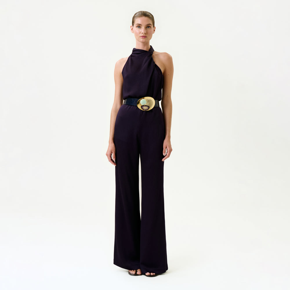 A woman poses against a plain white background wearing the Palermo Pant—an elegant, wide-leg, dark purple jumpsuit with an asymmetrical neckline and a bold belt featuring a gold circular buckle, crafted from luxurious Italian fabric.