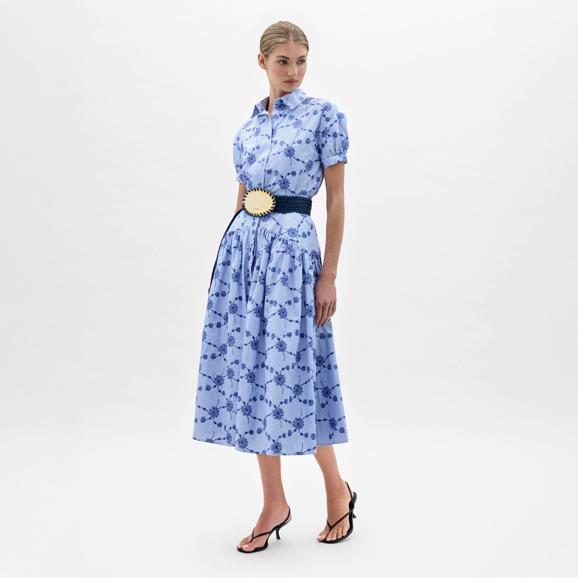 A woman wearing the Narel Skirt, styled with a light blue short-sleeve floral-print top, a wide dark belt, and black heeled sandals, stands against a plain white background.