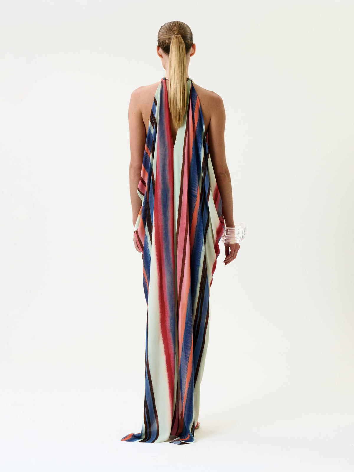 A person stands facing away, wearing the Niagara Dress—a long, backless design featuring vertical stripes in blue, red, and cream. Their hair is styled in a low ponytail, complemented by a large translucent bracelet. The background is plain white.