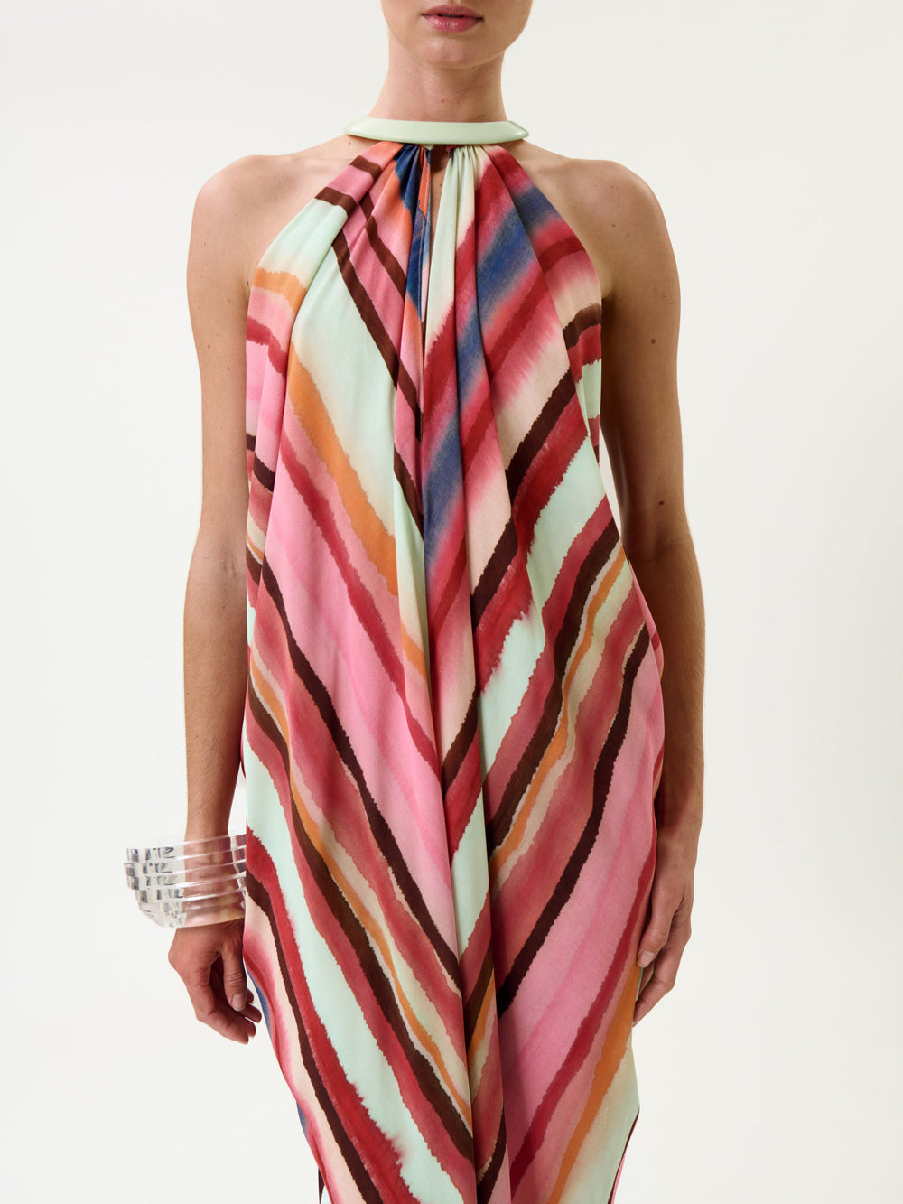 A person wearing the Niagara Dress—a sleeveless, halter-neck style featuring diagonal stripes in pink, red, blue, and mint green—along with clear chunky bracelets on their left wrist, stands against a plain white background.
