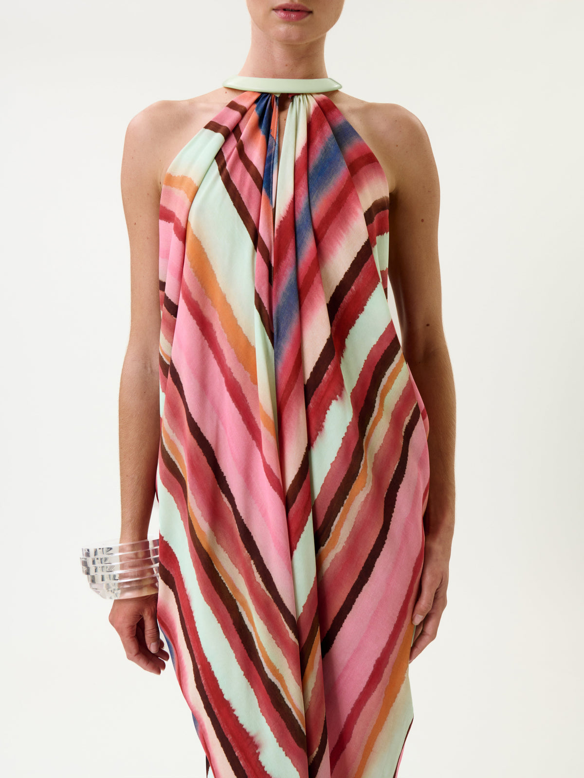 A person wearing the Niagara Dress—a sleeveless, halter-neck style featuring diagonal stripes in pink, red, blue, and mint green—along with clear chunky bracelets on their left wrist, stands against a plain white background.