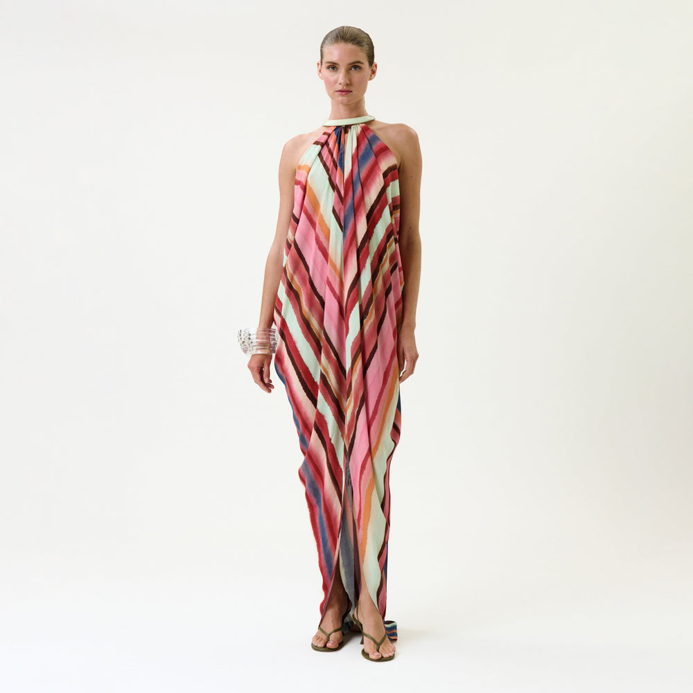 A woman wears the Niagara Dress, a sleeveless, floor-length gown featuring colorful diagonal stripes and a halter neckline, accessorized with chunky bracelets, standing against a plain white background.