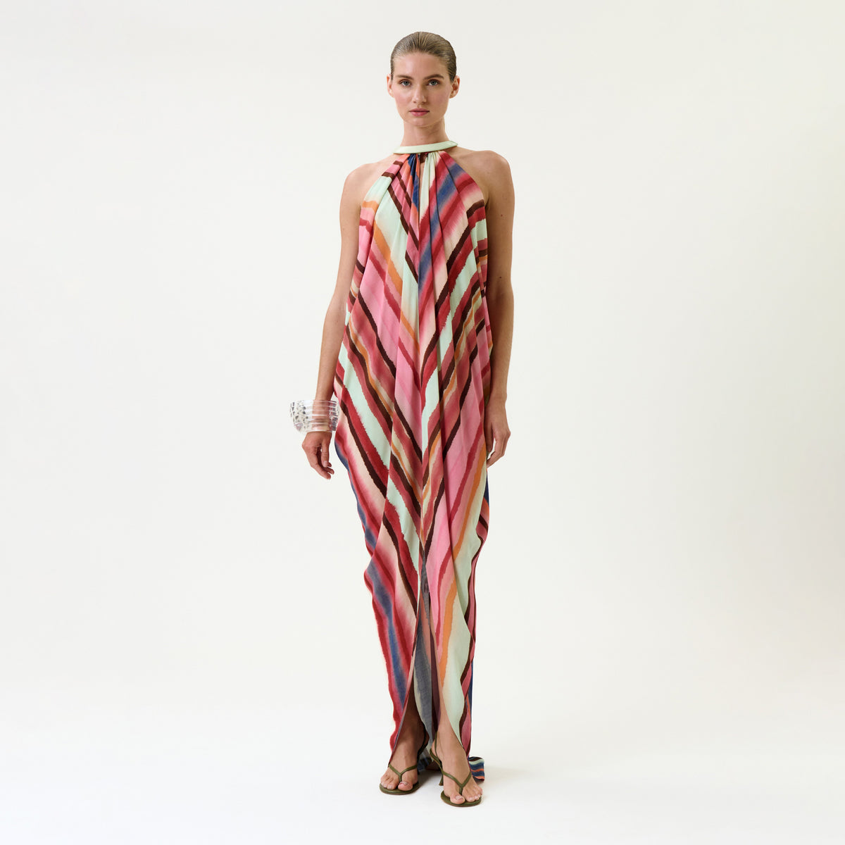 A woman wears the Niagara Dress, a sleeveless, floor-length gown featuring colorful diagonal stripes and a halter neckline, accessorized with chunky bracelets, standing against a plain white background.