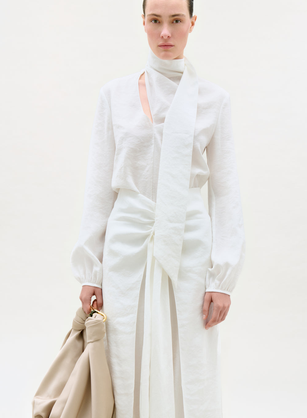 Woman wearing a white outfit holding a beige bag against a plain background