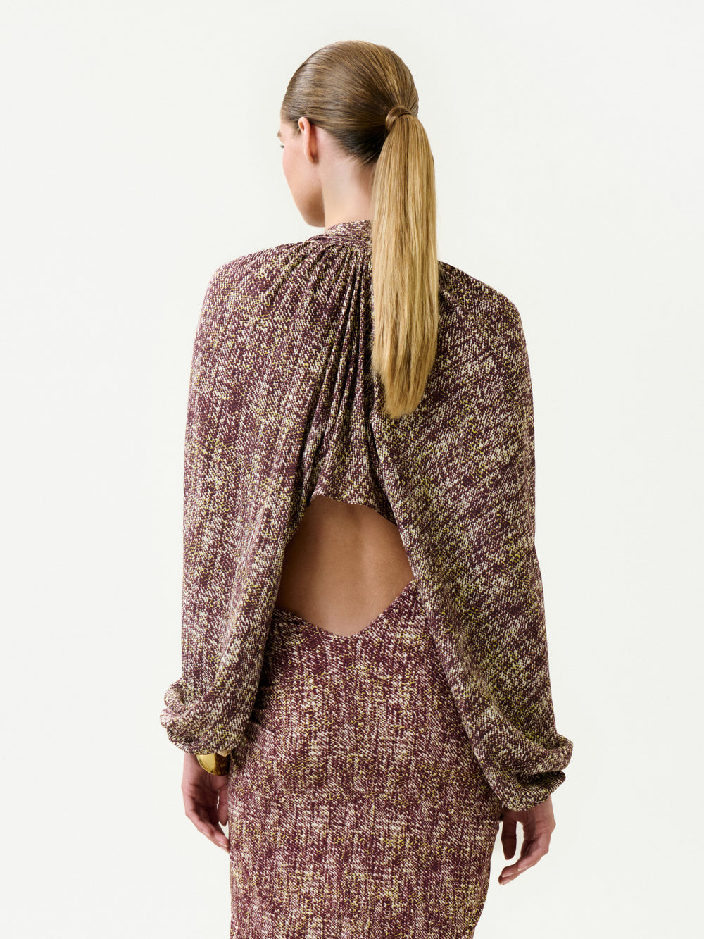 A person with long, straight blonde hair in a low ponytail wears the Marianela Cape, a textured multicolored dress with draped sleeves and an open back made from Italian fabric, standing against a plain white background.