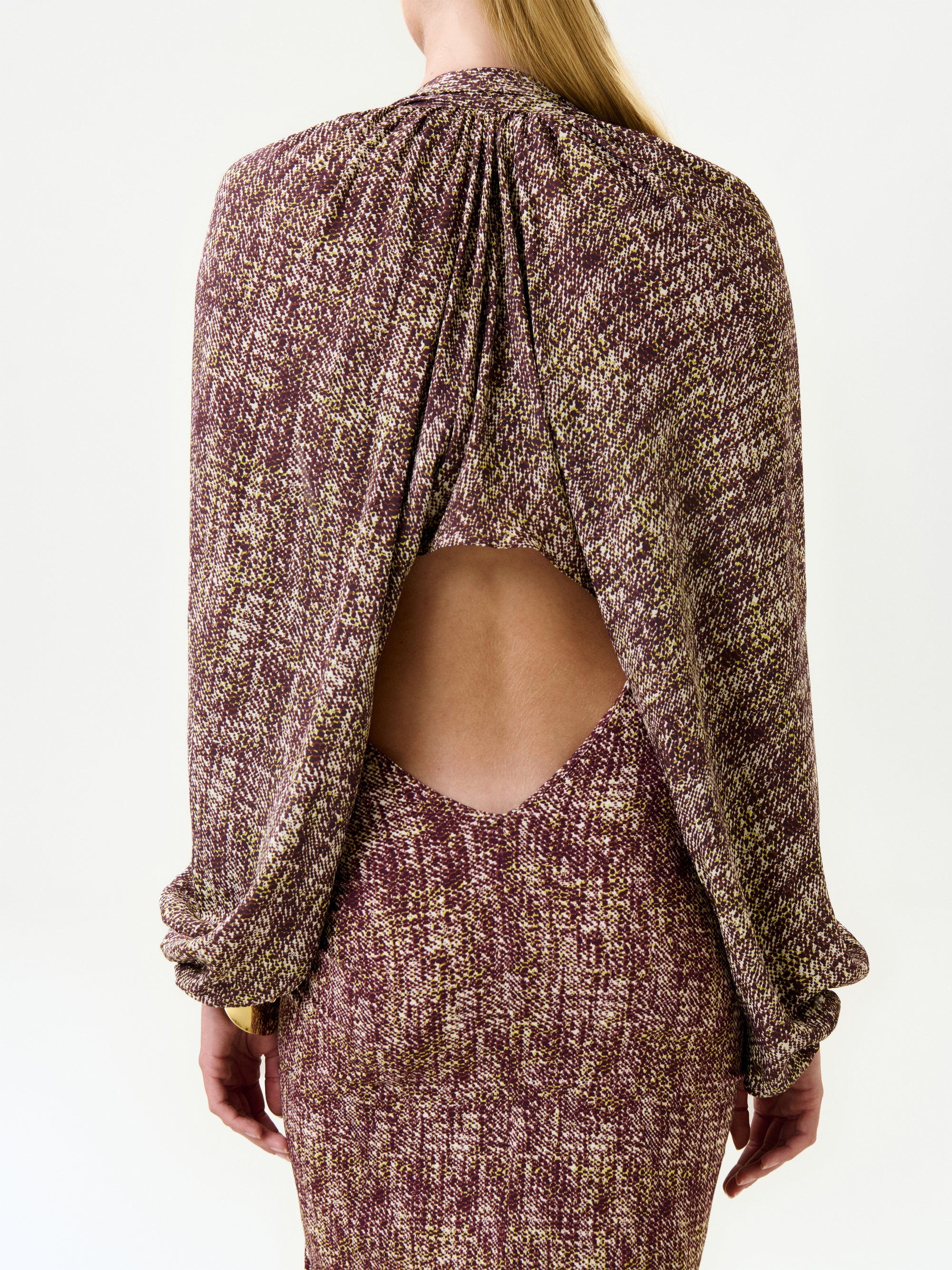 A person with long blonde hair wears the Marianela Cape, a textured, multicolored purple and beige dress made from Italian fabric with an open back and draped design, standing against a plain white background.