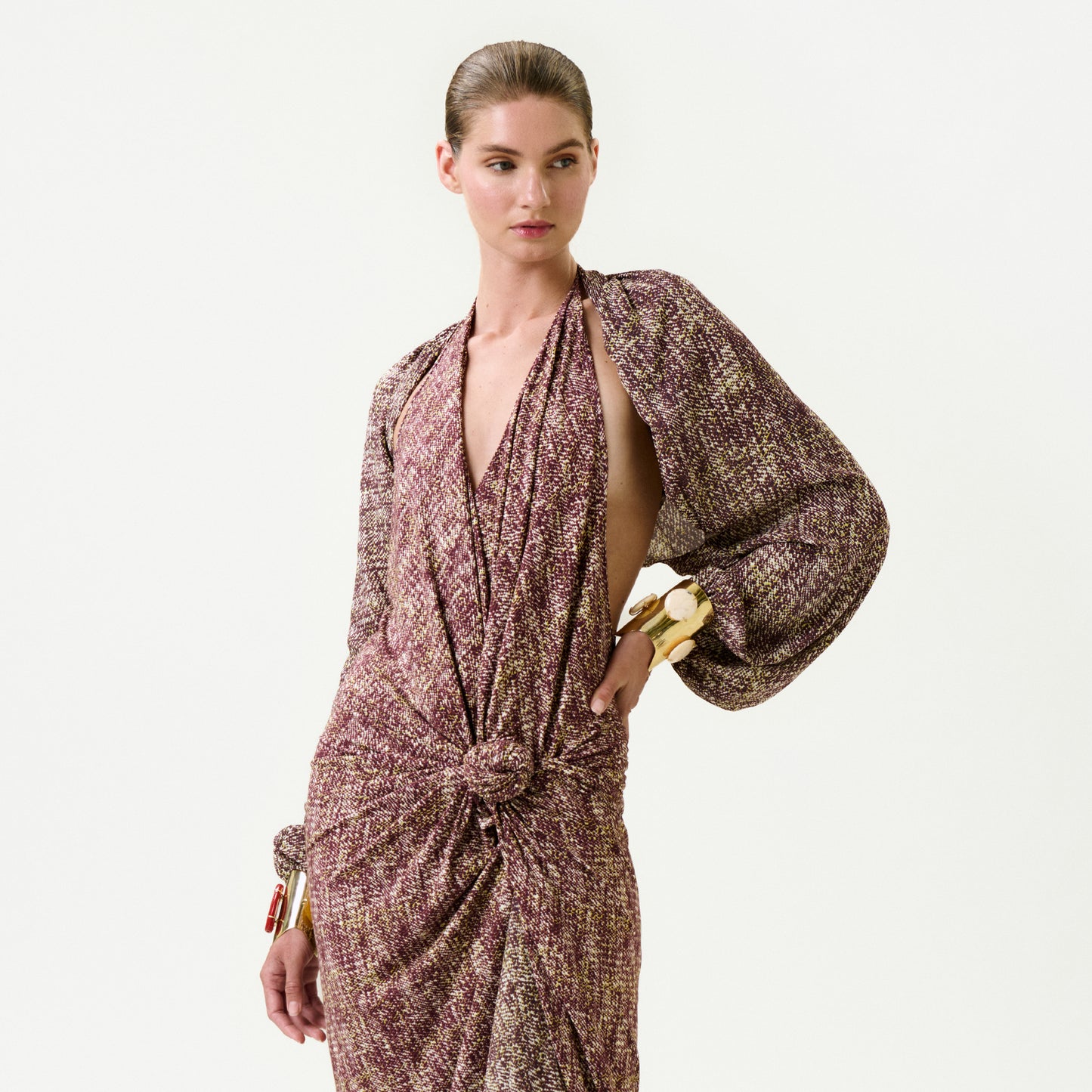 A woman models the Marianela Cape, a long-sleeved, textured purple and beige dress with a deep V-neckline and front knot, crafted from luxurious Italian fabric, styled with chunky gold bangles against a plain white backdrop.