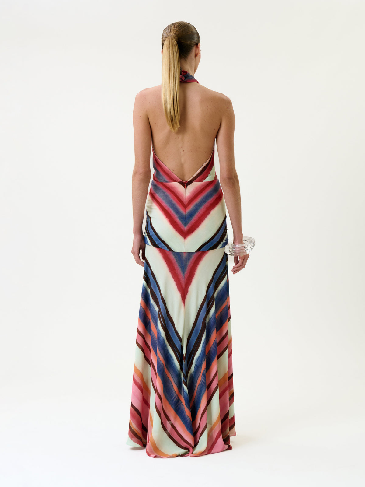 A person with long blonde hair in a ponytail wears the Patsy Dress—a backless halter design featuring colorful chevron stripes—paired with clear bangles, standing against a plain white background.