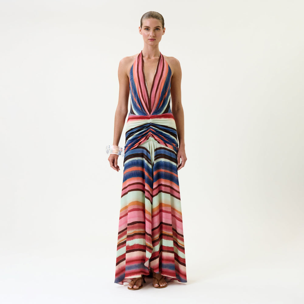 A woman wears the Patsy Dress—a sleeveless, halter-neck gown with a deep V-neckline and a multicolored striped pattern, featuring a knotted detail at the waist. She completes her look with sandals and bracelets, standing against a plain white background.