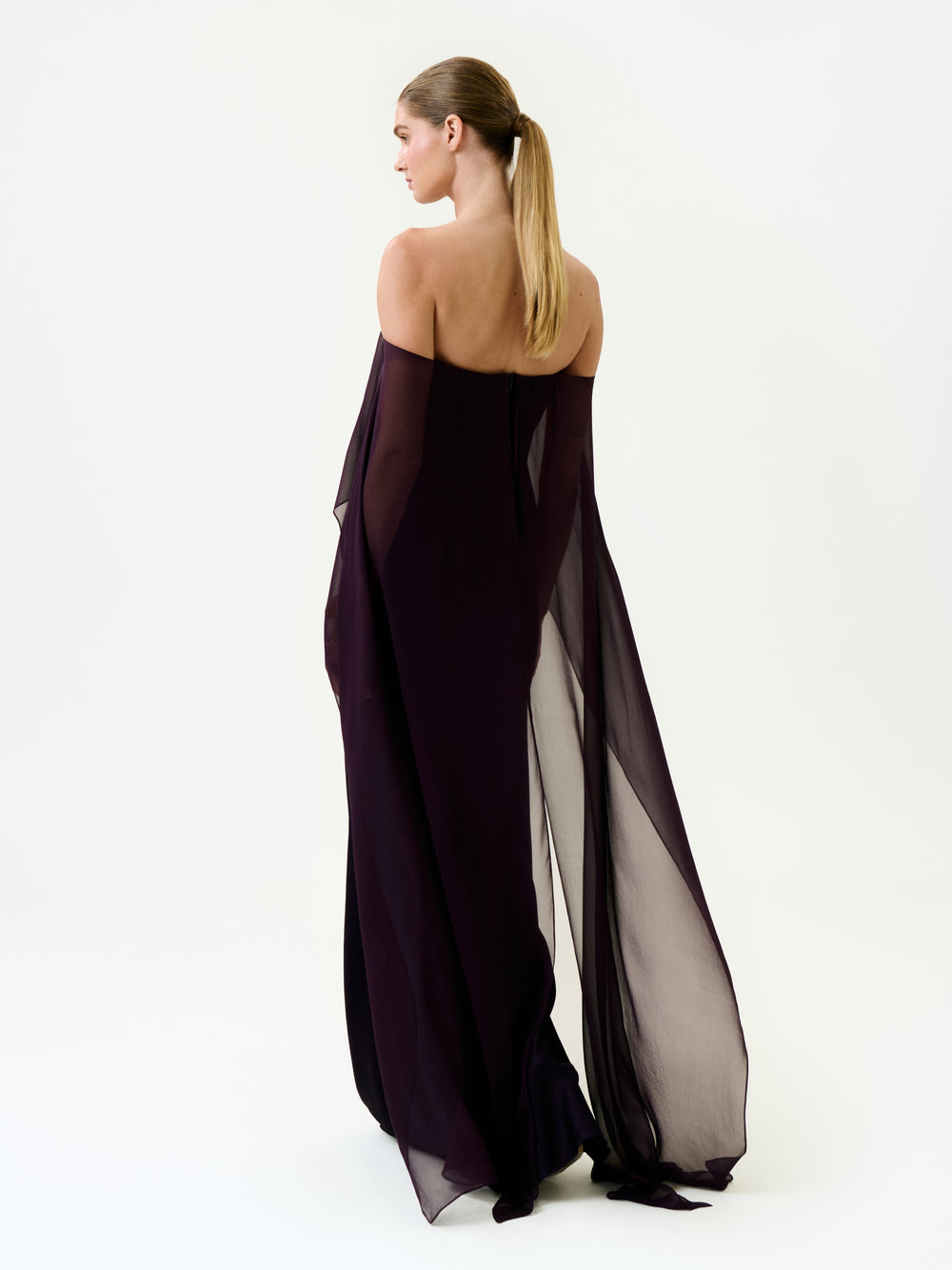 A woman with long, straight hair in a ponytail wears the Maiya Dress—an off-shoulder, floor-length sheer dark purple maxi made from Italian fabric—standing against a plain white background, facing away and slightly to the side.
