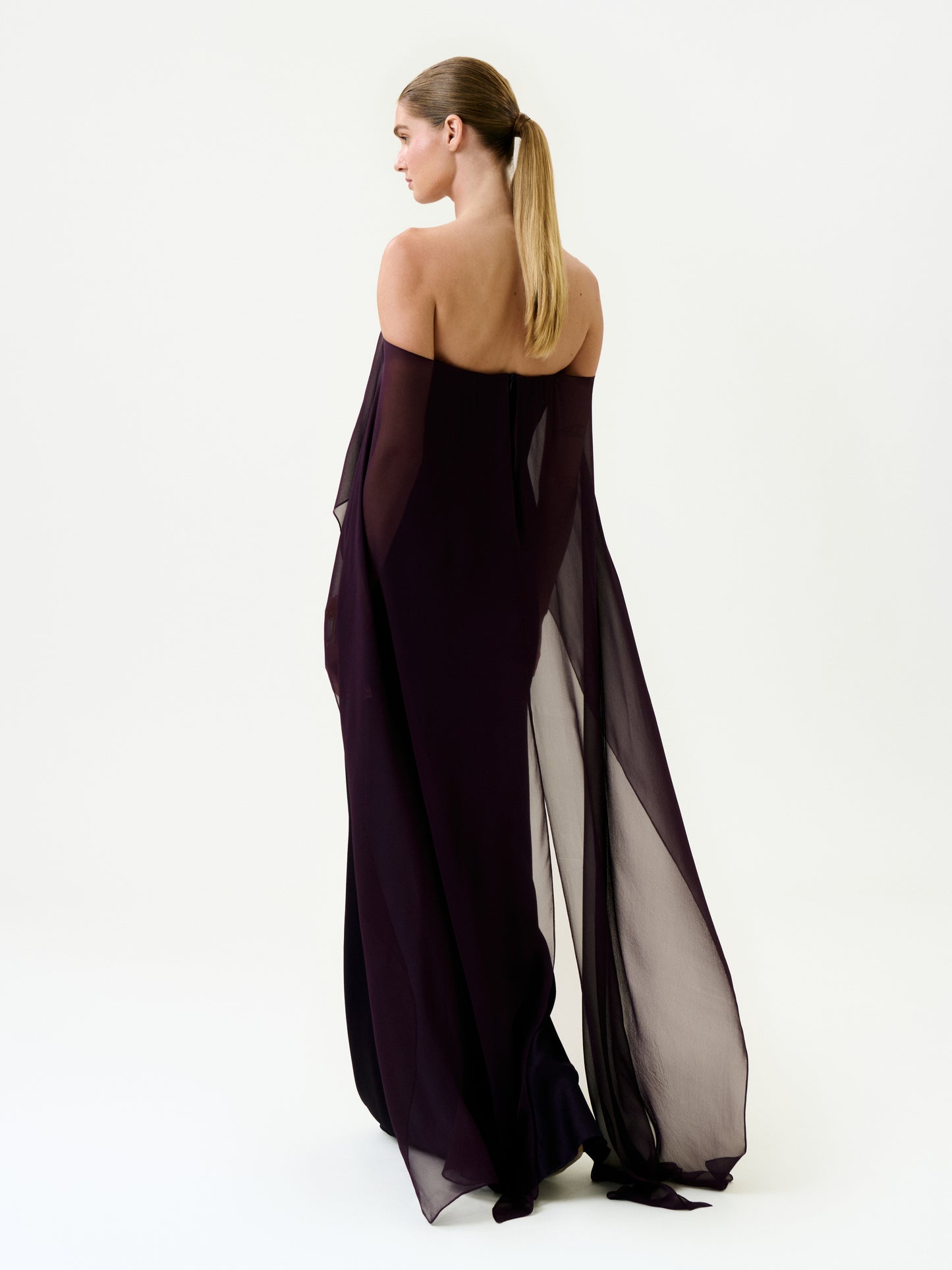 A woman with long, straight hair in a ponytail wears the Maiya Dress—an off-shoulder, floor-length sheer dark purple maxi made from Italian fabric—standing against a plain white background, facing away and slightly to the side.