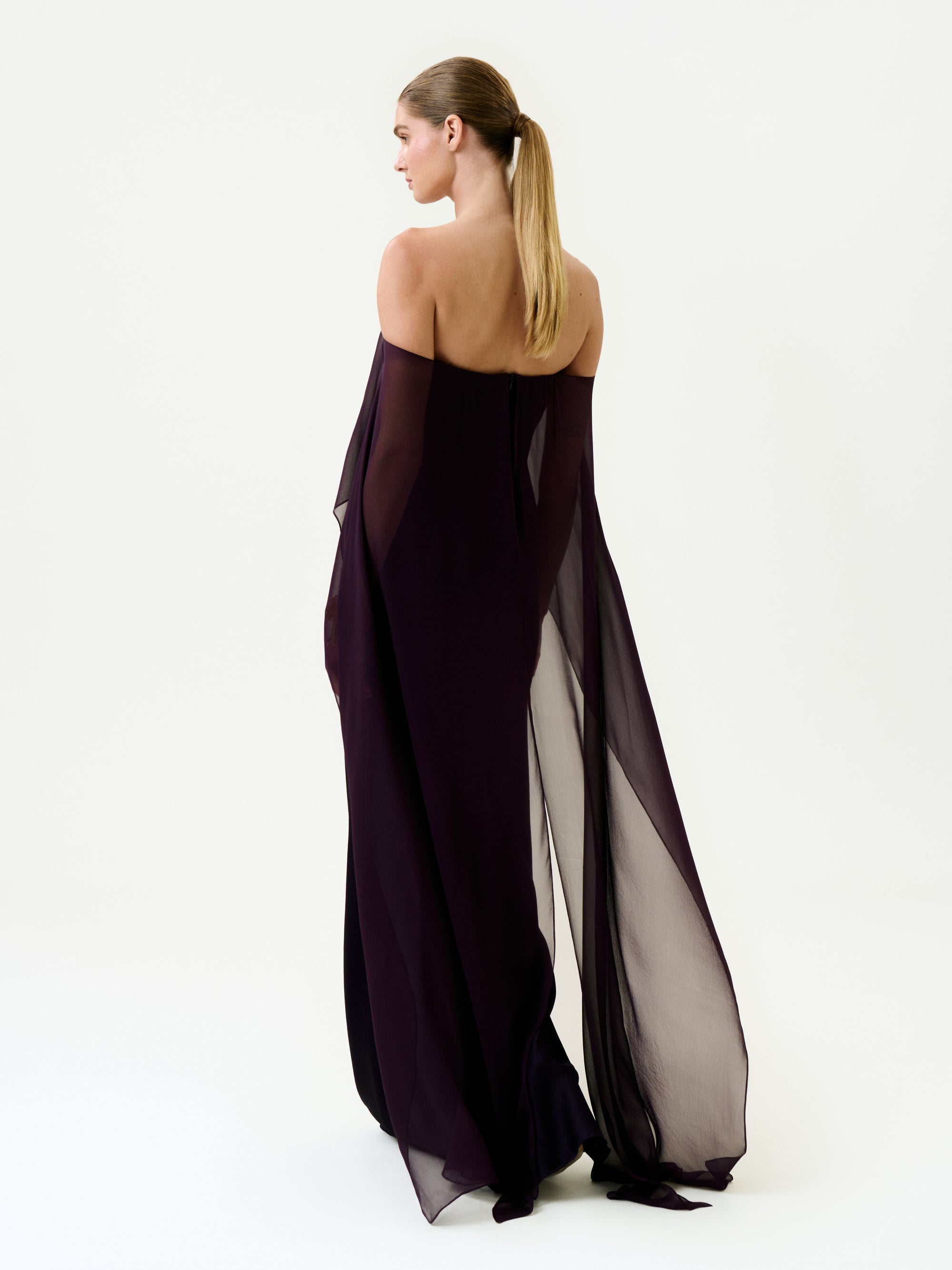 A woman with long, straight hair in a ponytail wears the Maiya Dress—an off-shoulder, floor-length sheer dark purple maxi made from Italian fabric—standing against a plain white background, facing away and slightly to the side.