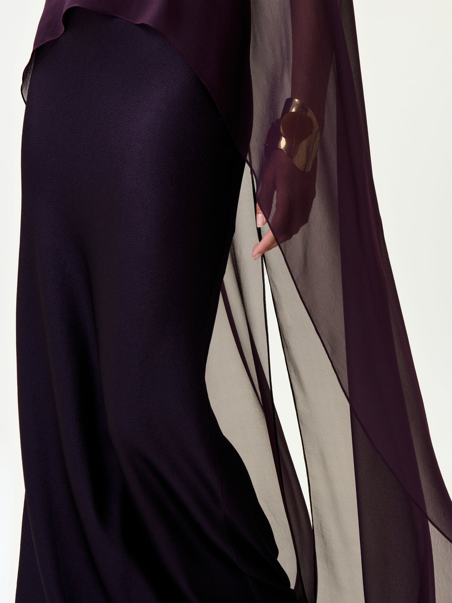 A person wears the Maiya Dress, a flowing sheer purple maxi crafted from luxurious Italian fabric, with a gold bracelet on their wrist. The dress drapes elegantly, highlighting its rich texture and graceful movement.