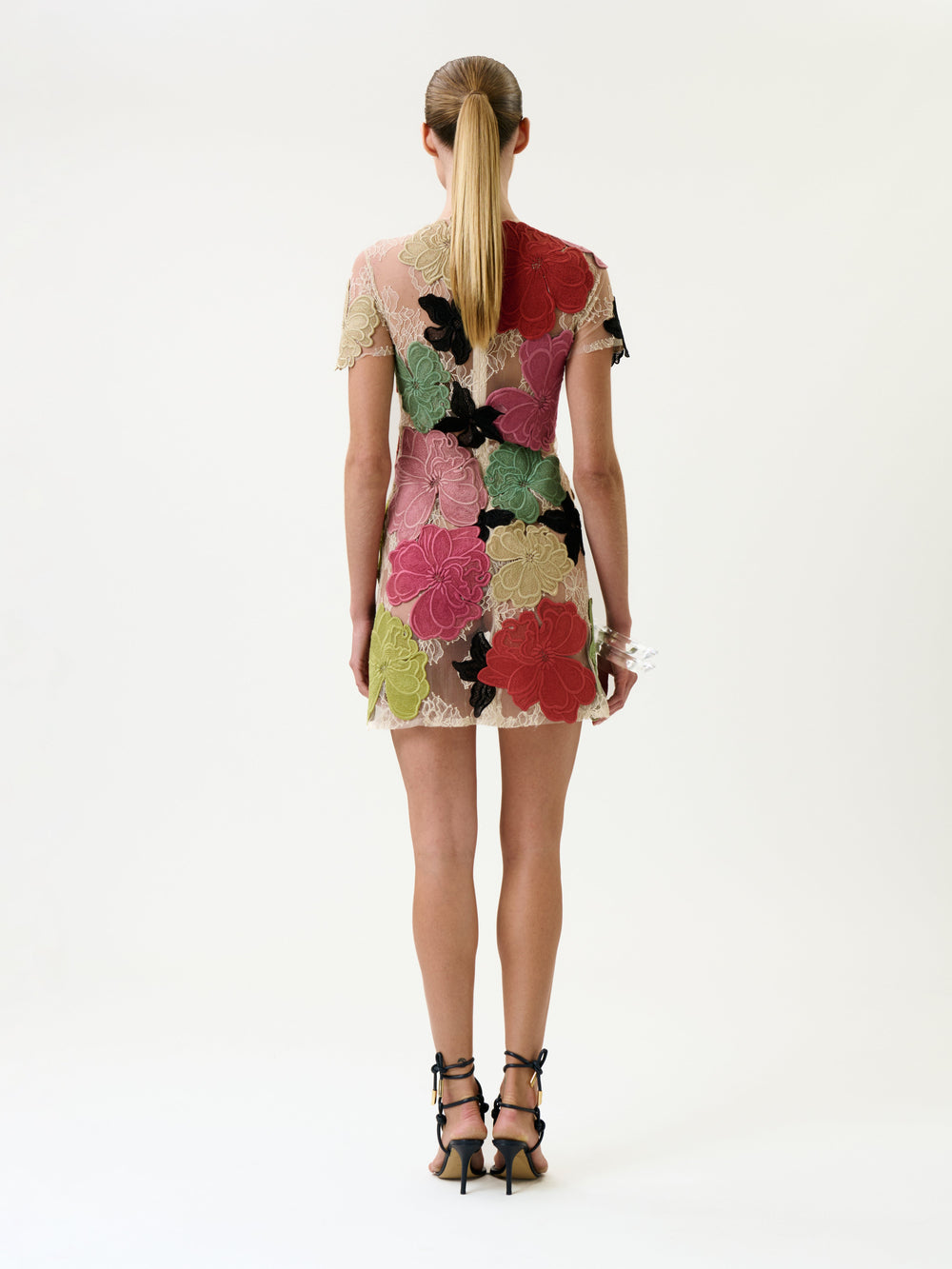 A woman with a low ponytail stands facing away, wearing the Hada Dress—a short, sheer dress adorned with large colorful floral patterns—paired with black strappy heels, against a plain white background.