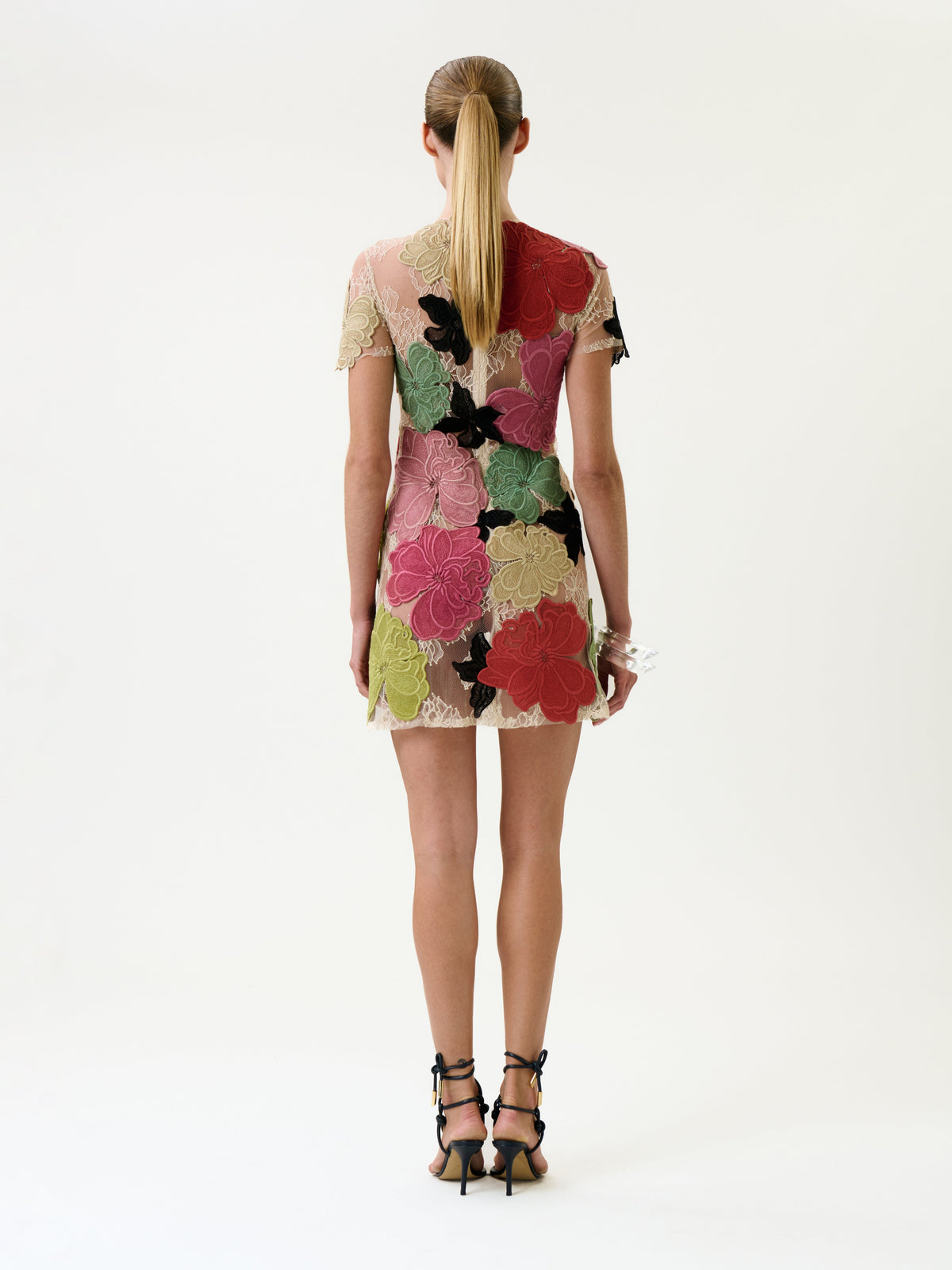A woman with a low ponytail stands facing away, wearing the Hada Dress—a short, sheer dress adorned with large colorful floral patterns—paired with black strappy heels, against a plain white background.