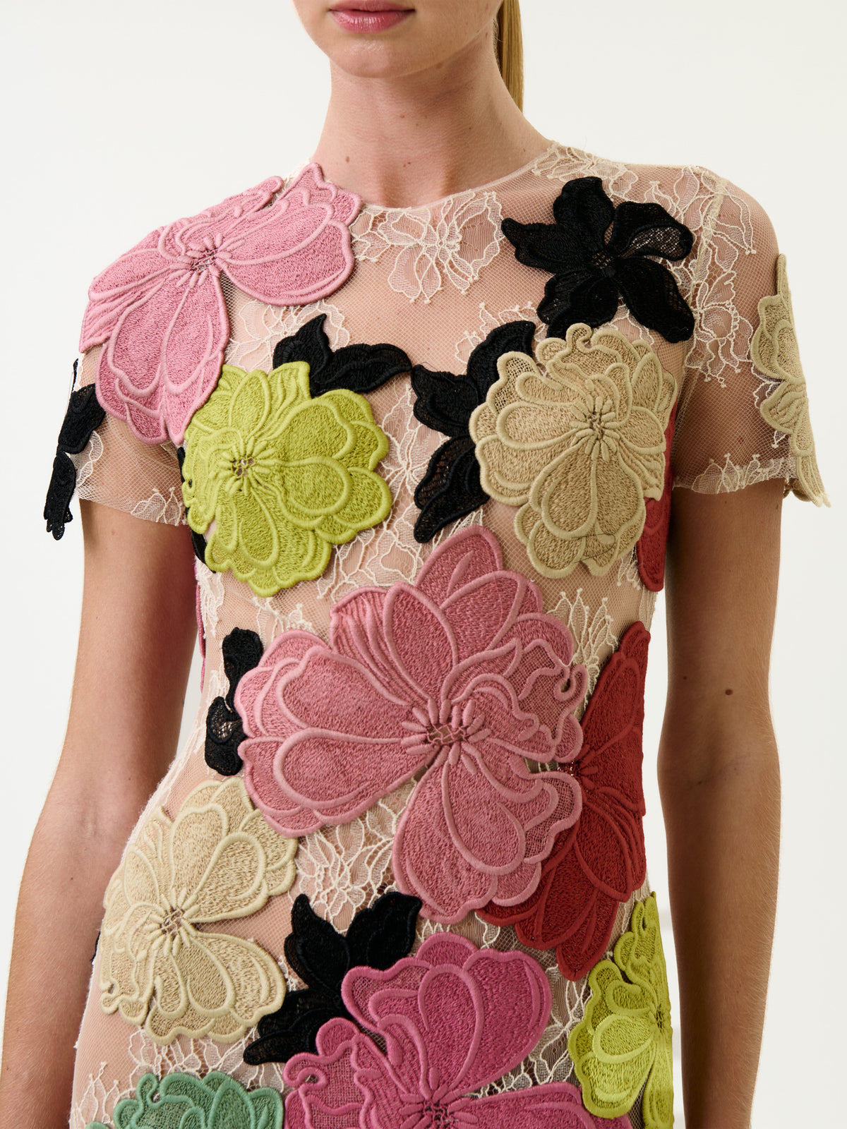 A woman wearing the Hada Dress, a sheer garment featuring large, colorful embroidered floral appliqués in pink, green, beige, and black. The dress has short sleeves and is finished with intricate lace details.
