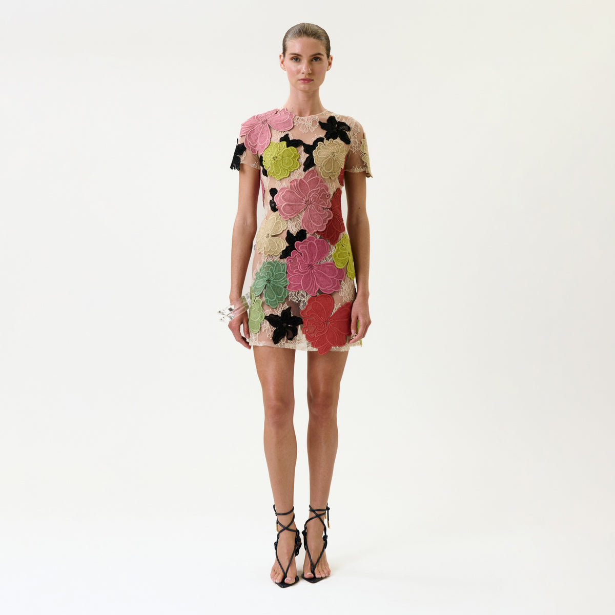 A woman stands facing forward in the Hada Dress, a short, fitted dress featuring large, colorful floral patterns. She pairs it with black strappy high heels and poses against a plain white background.
