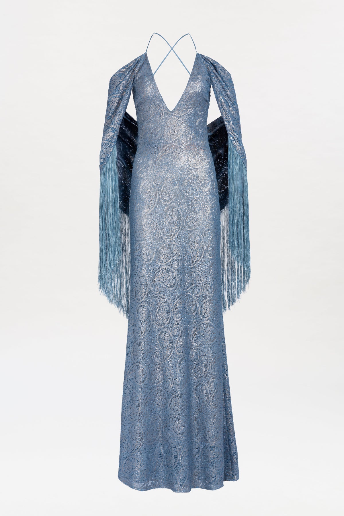 The Habana Dress is a shimmering blue, floor-length gown featuring a deep V-neckline, delicate crisscross straps, and long fringed shawl sleeves. This elegant dress showcases a subtle paisley pattern and a slim, sophisticated silhouette.