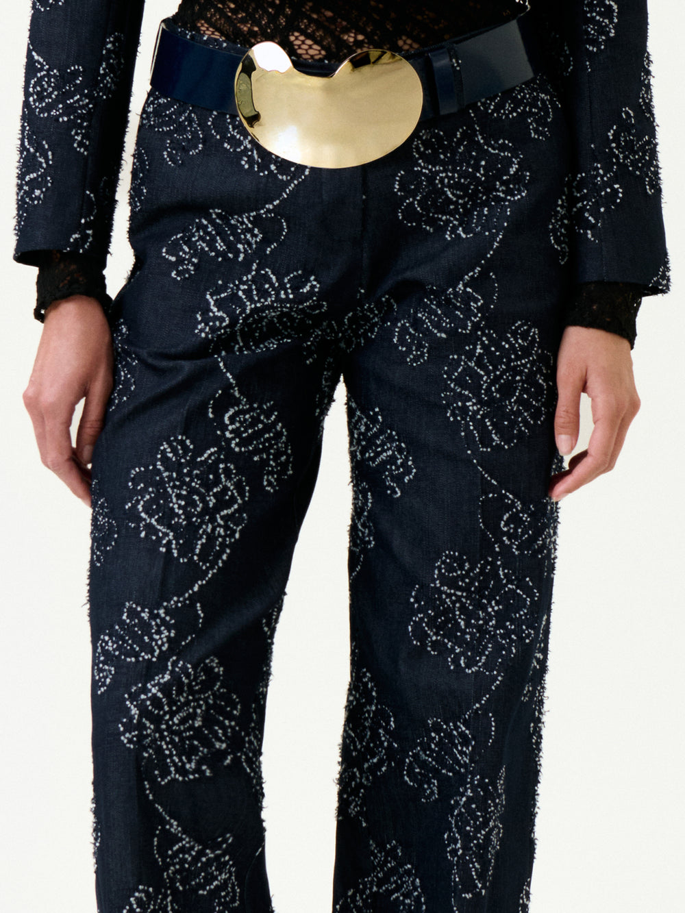 A person wears the Ulla Pant, featuring dark floral embroidery, paired with a black lace top and a wide belt with a gold oval buckle. The true-to-size fit adds chic appeal. Image shows them cropped from shoulders to knees.