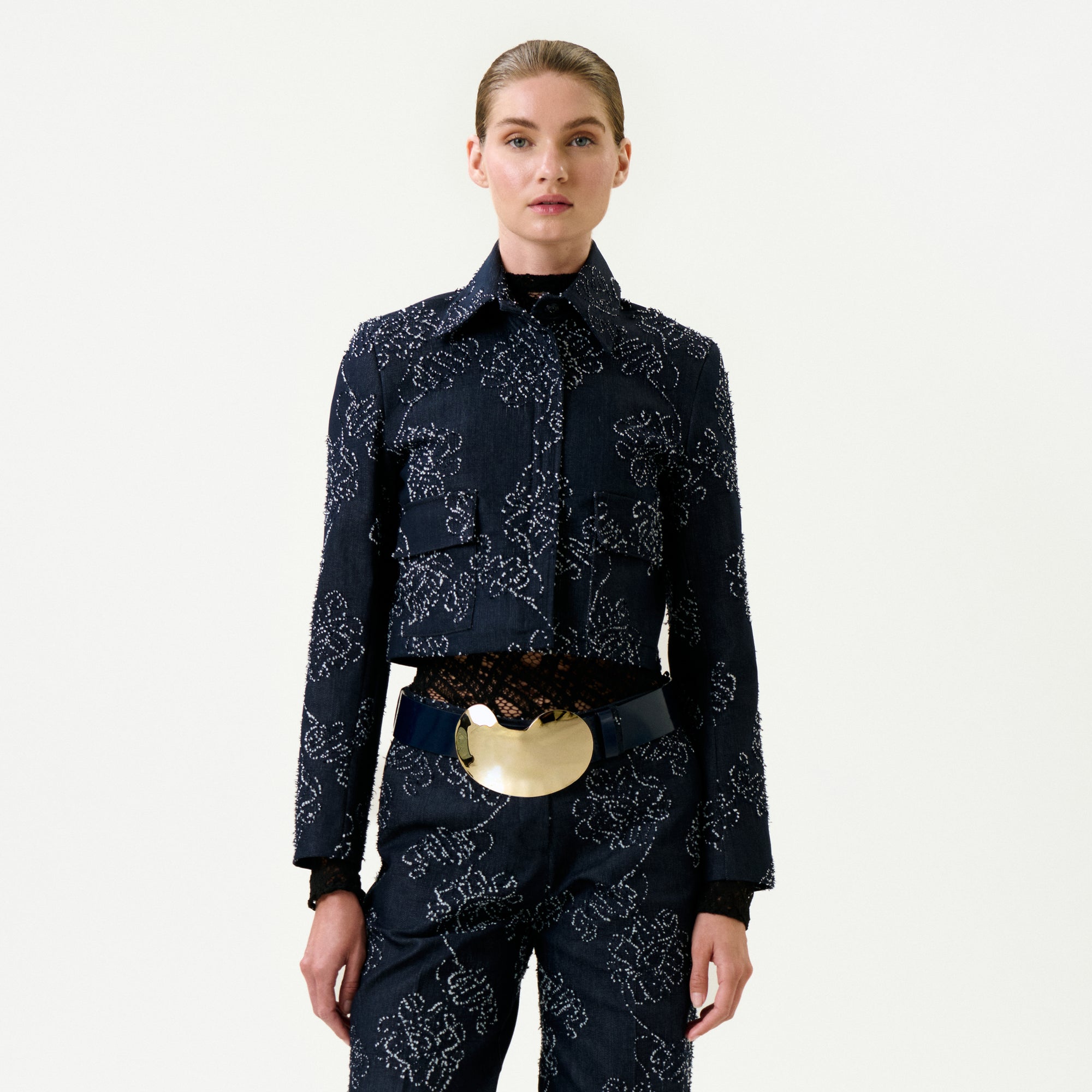 A woman stands facing forward in the Zulma Jacket, crafted from dark blue Italian fabric with matching pants. Her slicked-back hair and bold gold belt buckle accentuate the true-to-size fit against a plain white background.