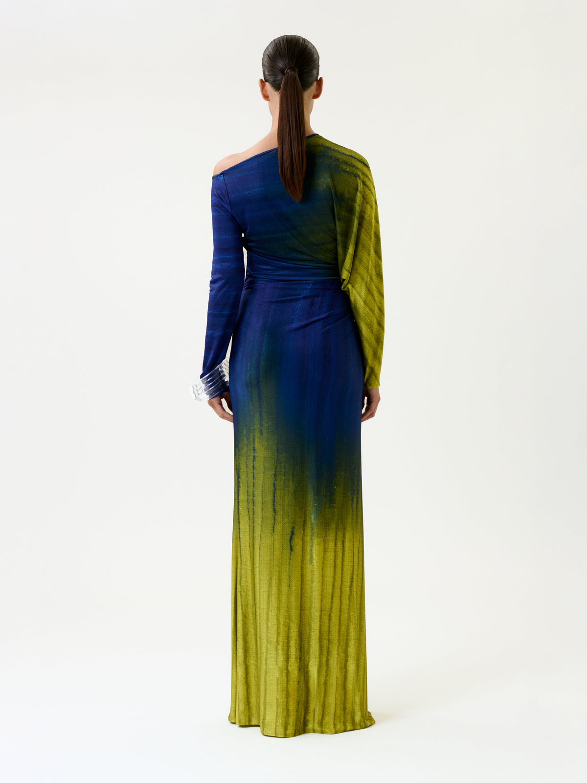 A woman with straight brown hair in a ponytail models the Saree Dress, a long, one-shoulder gown featuring a blue and yellow gradient, as she stands facing away against a plain light background.