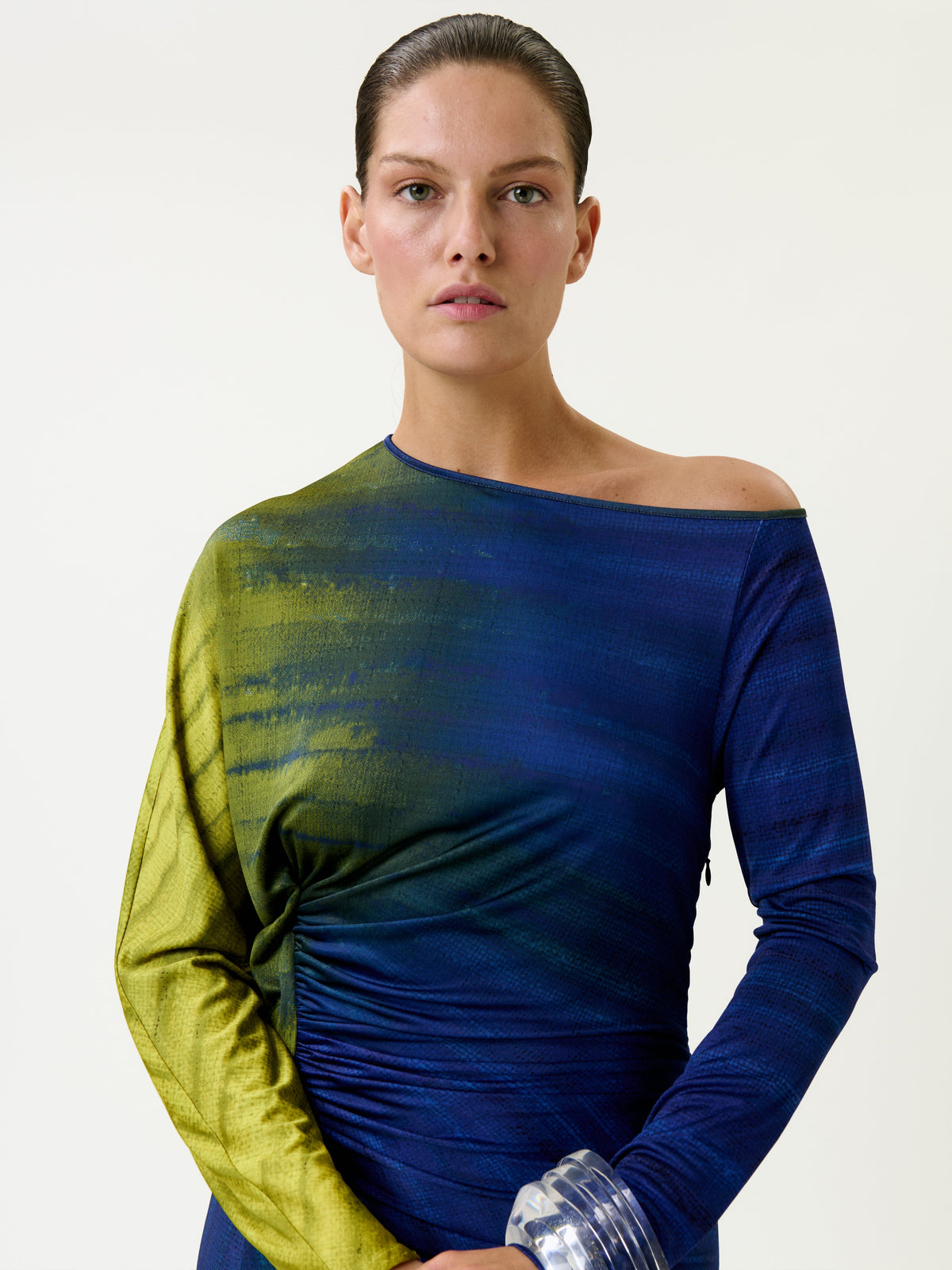 A person with slicked-back hair wears the Saree Dress, featuring blue and green tones and a one-shoulder design, while holding a large clear bracelet. The background is plain and light-colored.