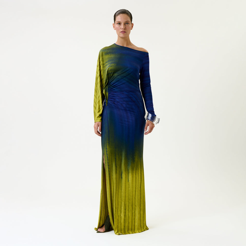 A woman stands against a plain background wearing the Saree Dress, a long, one-shoulder gown featuring blue and yellow ombre coloring and a side slit.