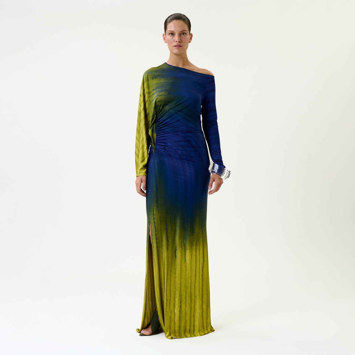 A woman stands against a plain background wearing the Saree Dress, a long, one-shoulder gown featuring blue and yellow ombre coloring and a side slit.