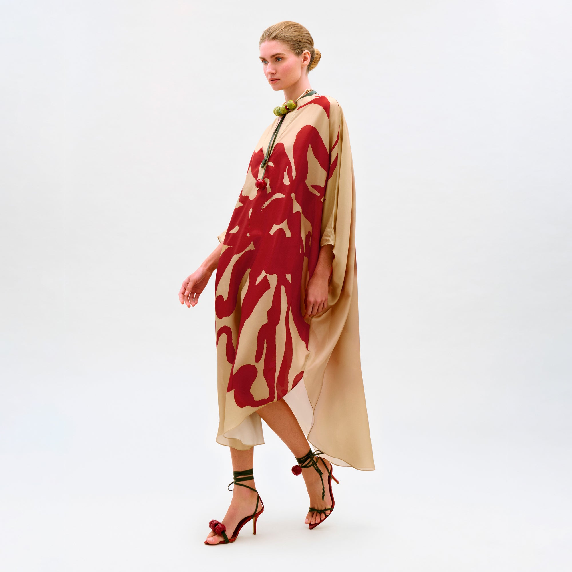 A woman in the Elea Tunic—a loose, beige 100% silk maxi dress with bold red abstract patterns—paired with a green beaded necklace and strappy red heels walks against a plain white background.
