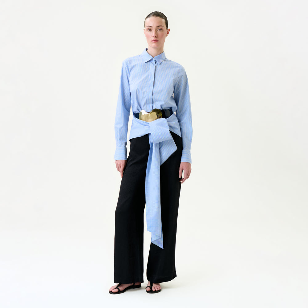 A person stands against a plain white background wearing the Ulla Pant in black wide-leg style, paired with a light blue button-up shirt and a large blue and beige Italian fabric belt with an oversized bow at the waist.