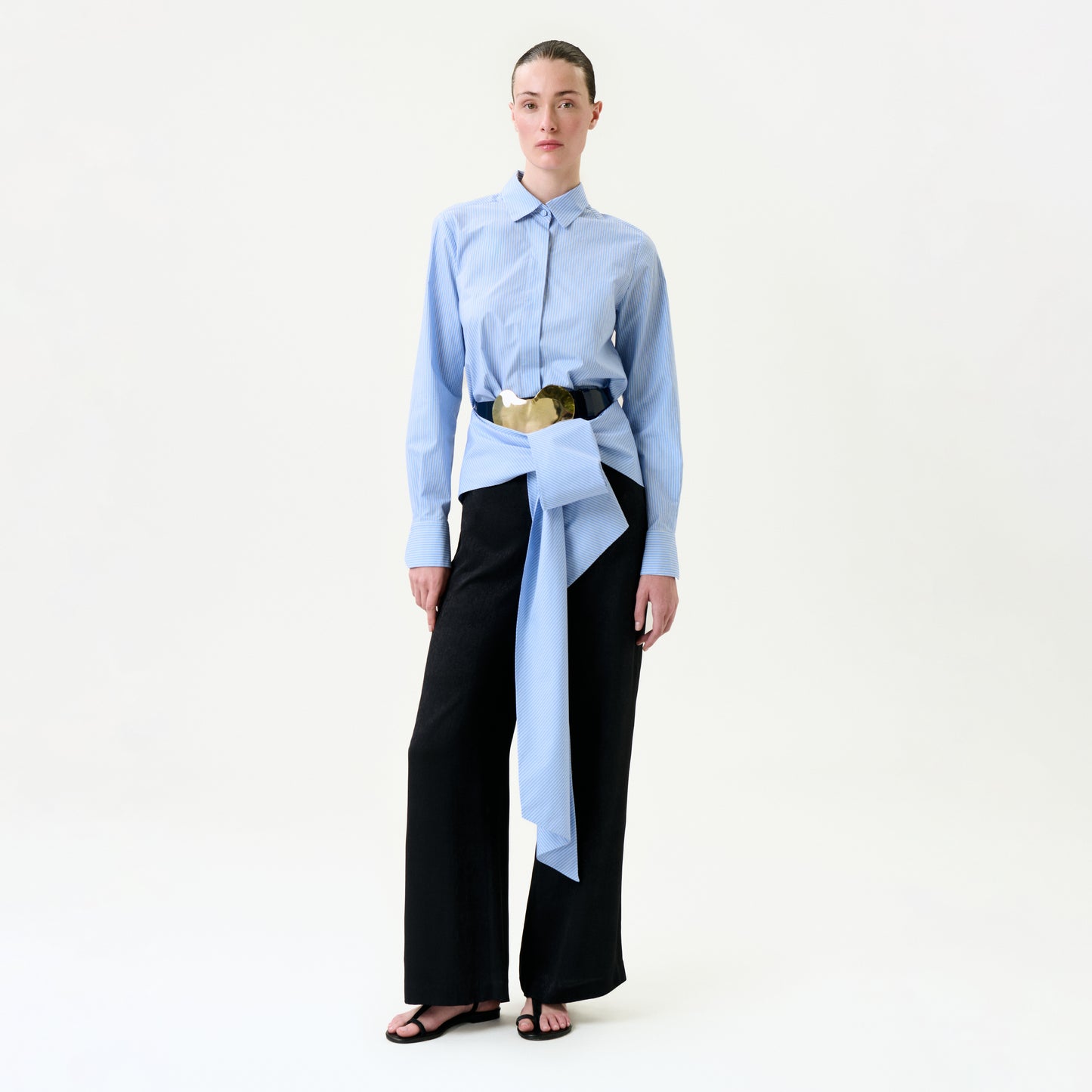 A person stands against a plain white background wearing the Ulla Pant in black wide-leg style, paired with a light blue button-up shirt and a large blue and beige Italian fabric belt with an oversized bow at the waist.