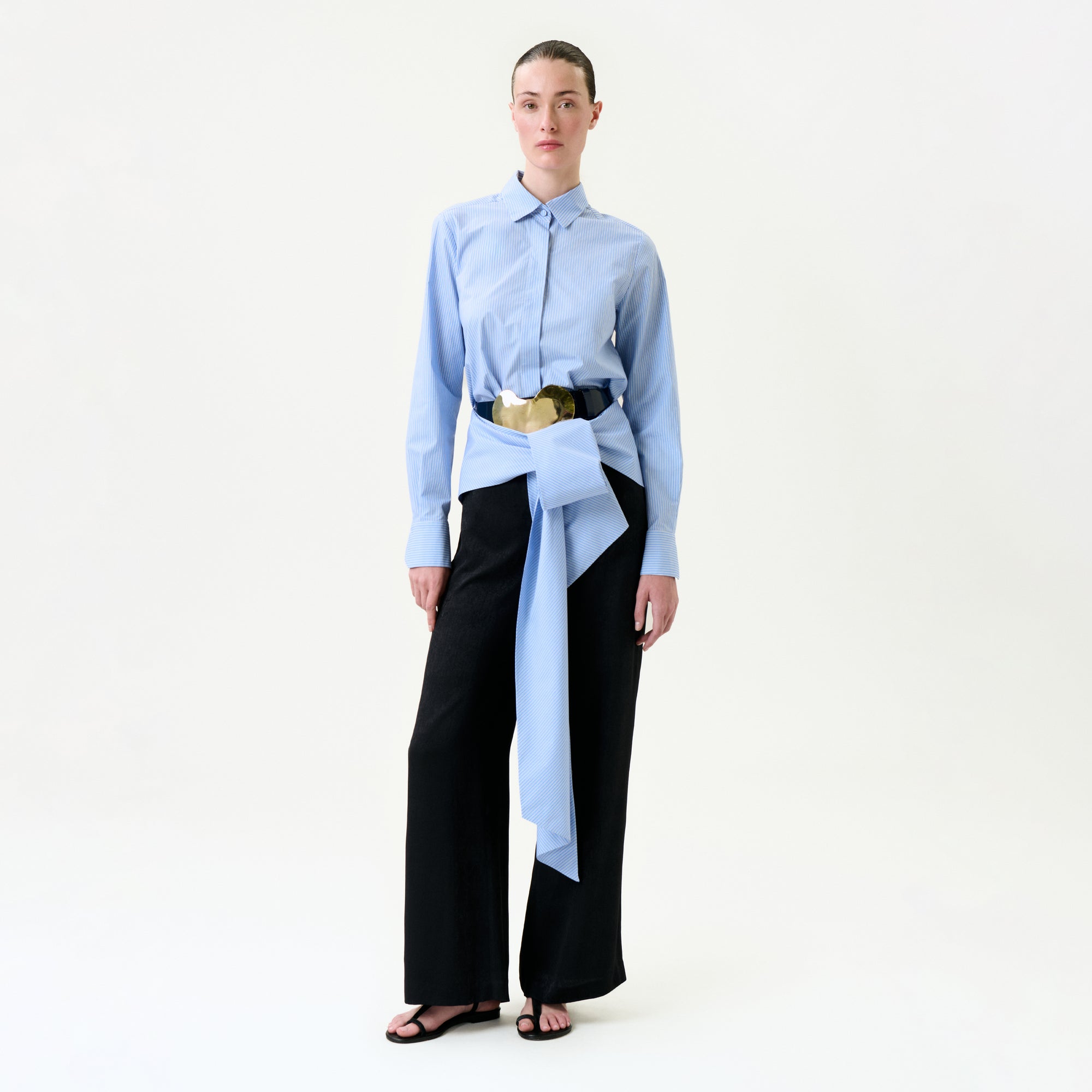 A person stands against a plain white background wearing the Ulla Pant in black wide-leg style, paired with a light blue button-up shirt and a large blue and beige Italian fabric belt with an oversized bow at the waist.