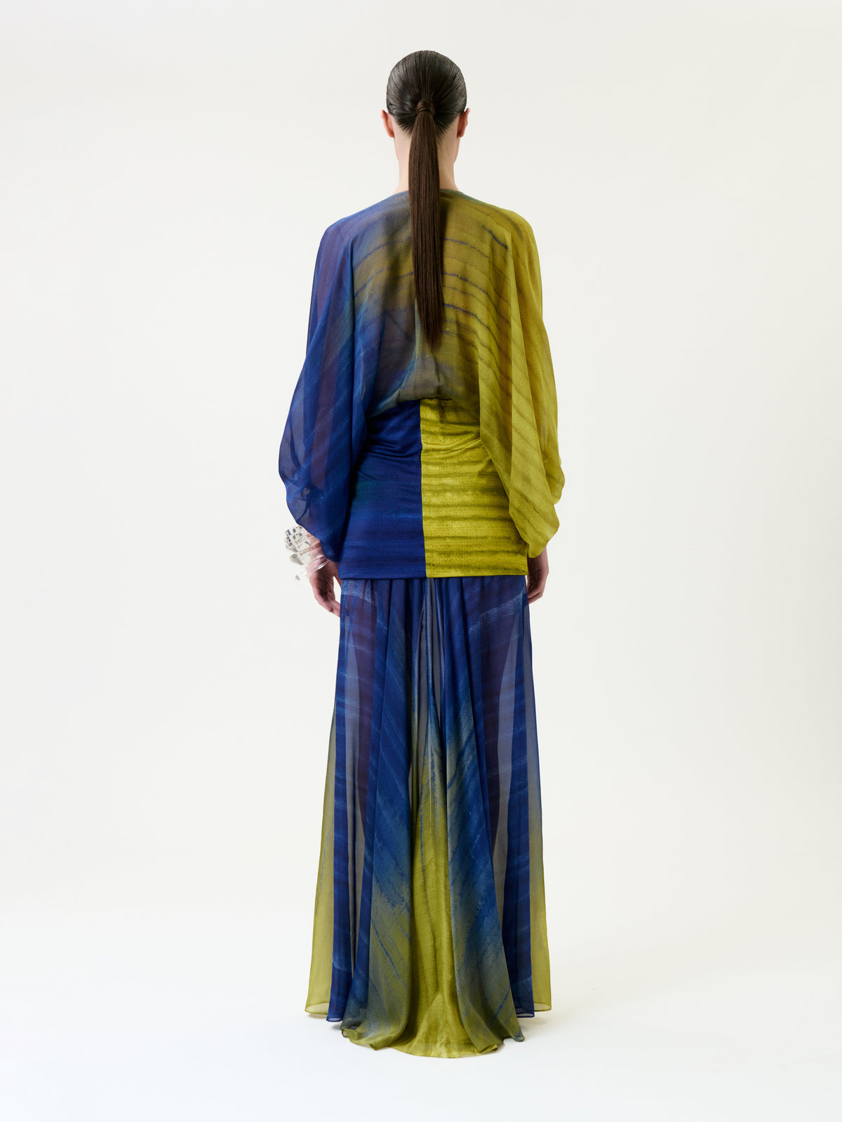 A person with long dark hair styled in a low ponytail is seen from the back wearing the Lilen Dress, a sheer, flowing blue and yellow piece, standing against a plain white background.