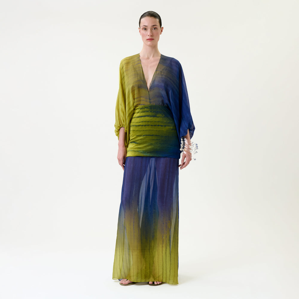A woman stands facing forward on a plain white background, wearing the Lilen Dress—a flowing, long dress featuring a deep V-neck and blue-to-yellow gradient hues.