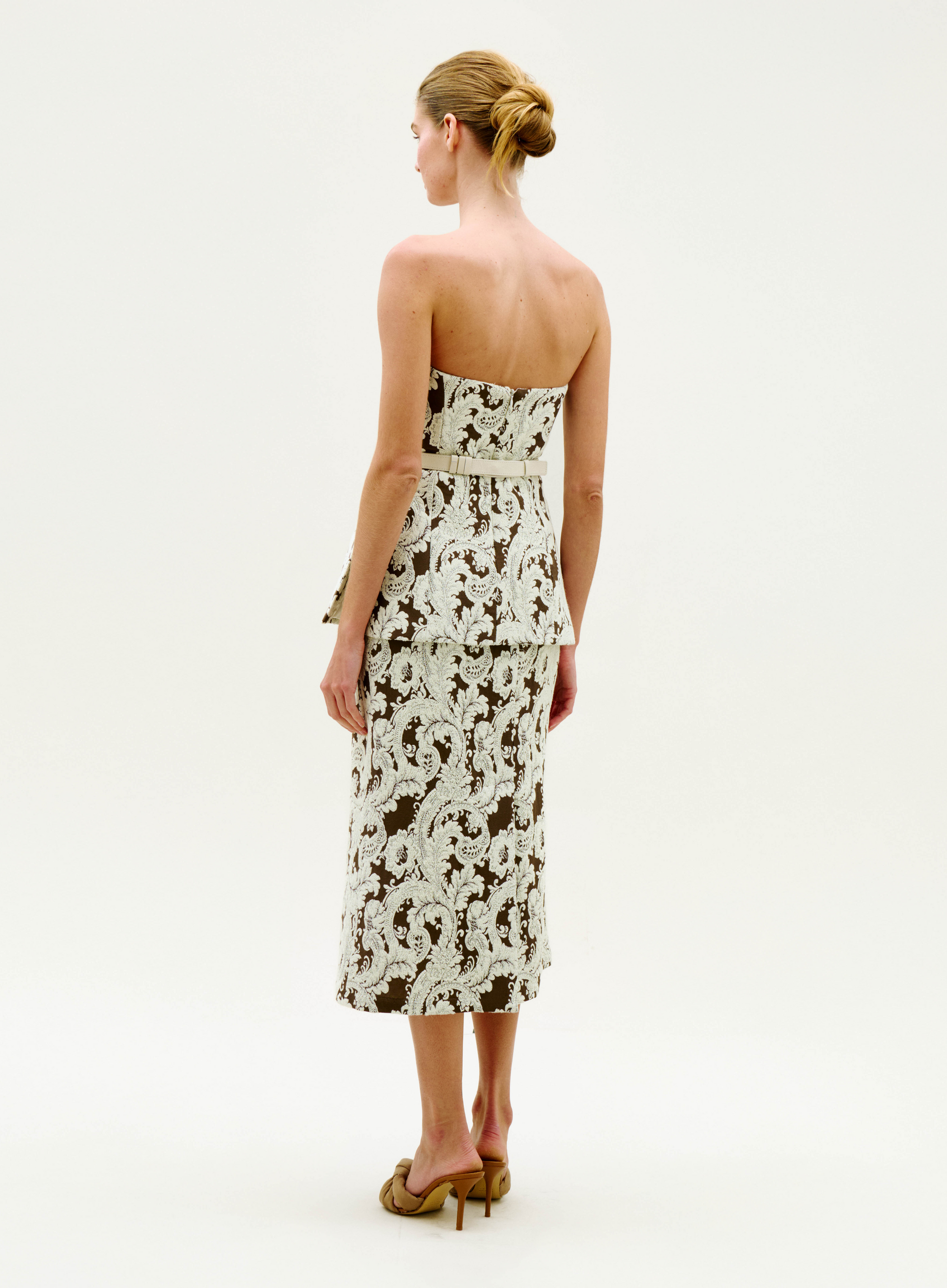 A woman faces away, wearing the Atira Skirt—a fitted piece with a white floral lace pattern over a dark base—and tan high heels. Her hair is in a neat bun, set against a plain light background.