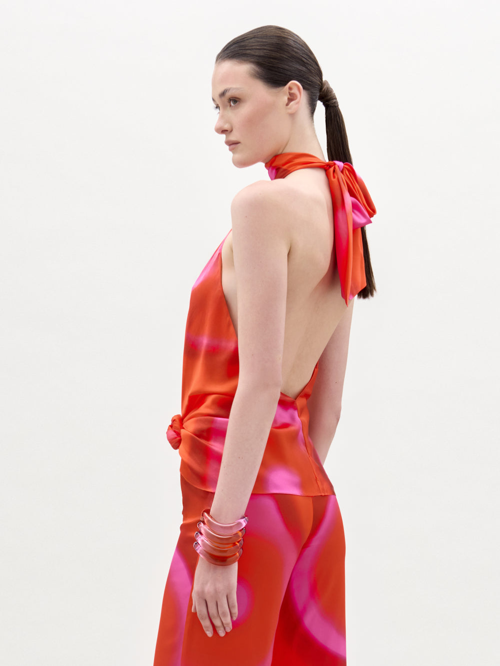 A woman with straight brown hair in a low ponytail models the Dita Blouse, a bright red and pink silk halter top with bold swirls and an open back, paired with matching pants and stacked pink bracelets on her wrist.