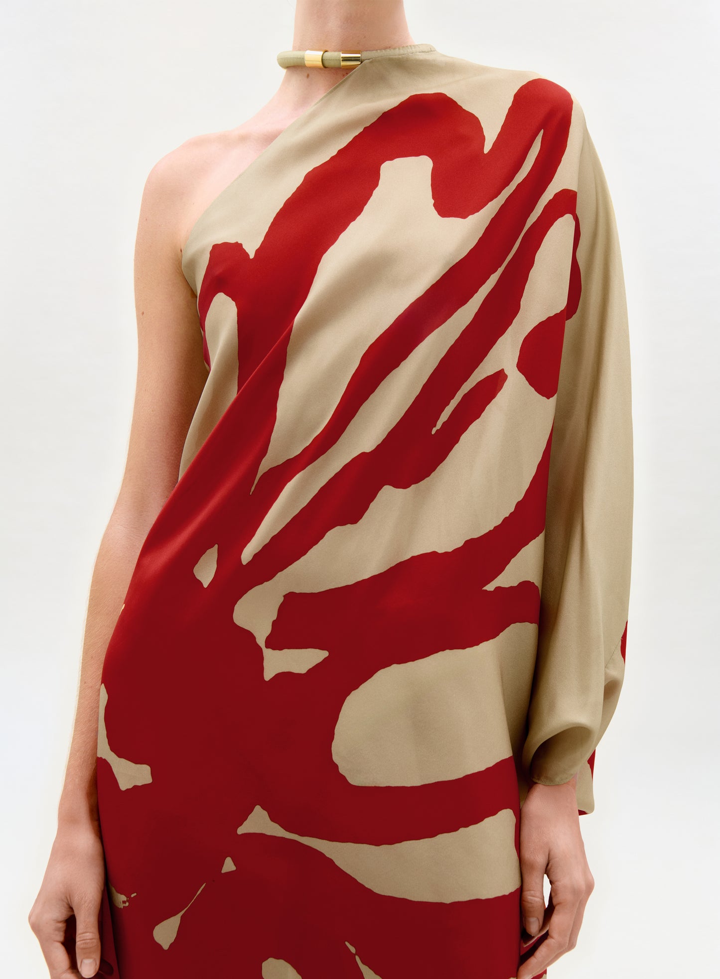 Someone models the Erica Dress—a tan one-shoulder silk maxi with a bold red abstract print and a single long sleeve—paired with a gold choker, set against a simple light background.