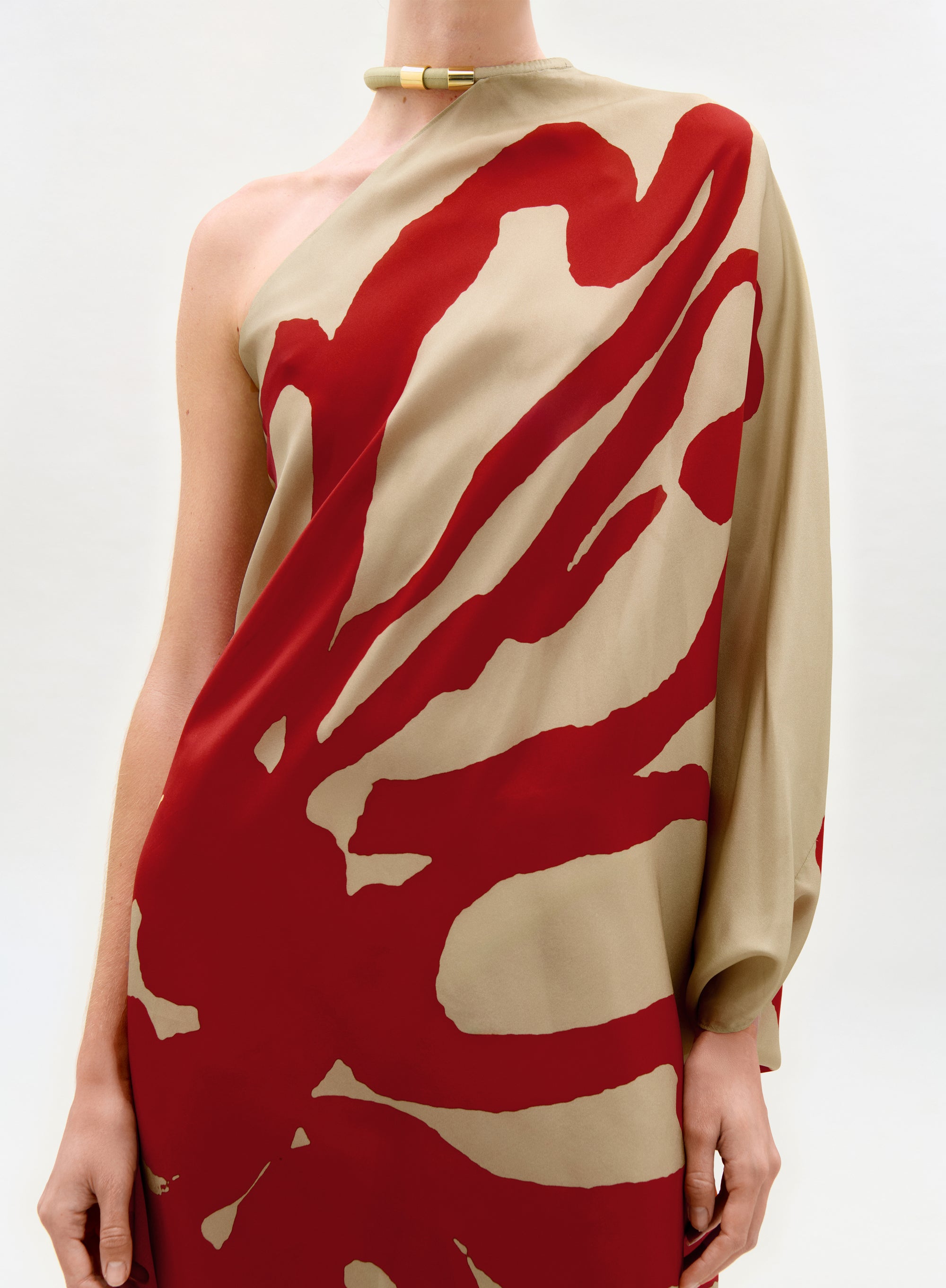 Someone models the Erica Dress—a tan one-shoulder silk maxi with a bold red abstract print and a single long sleeve—paired with a gold choker, set against a simple light background.