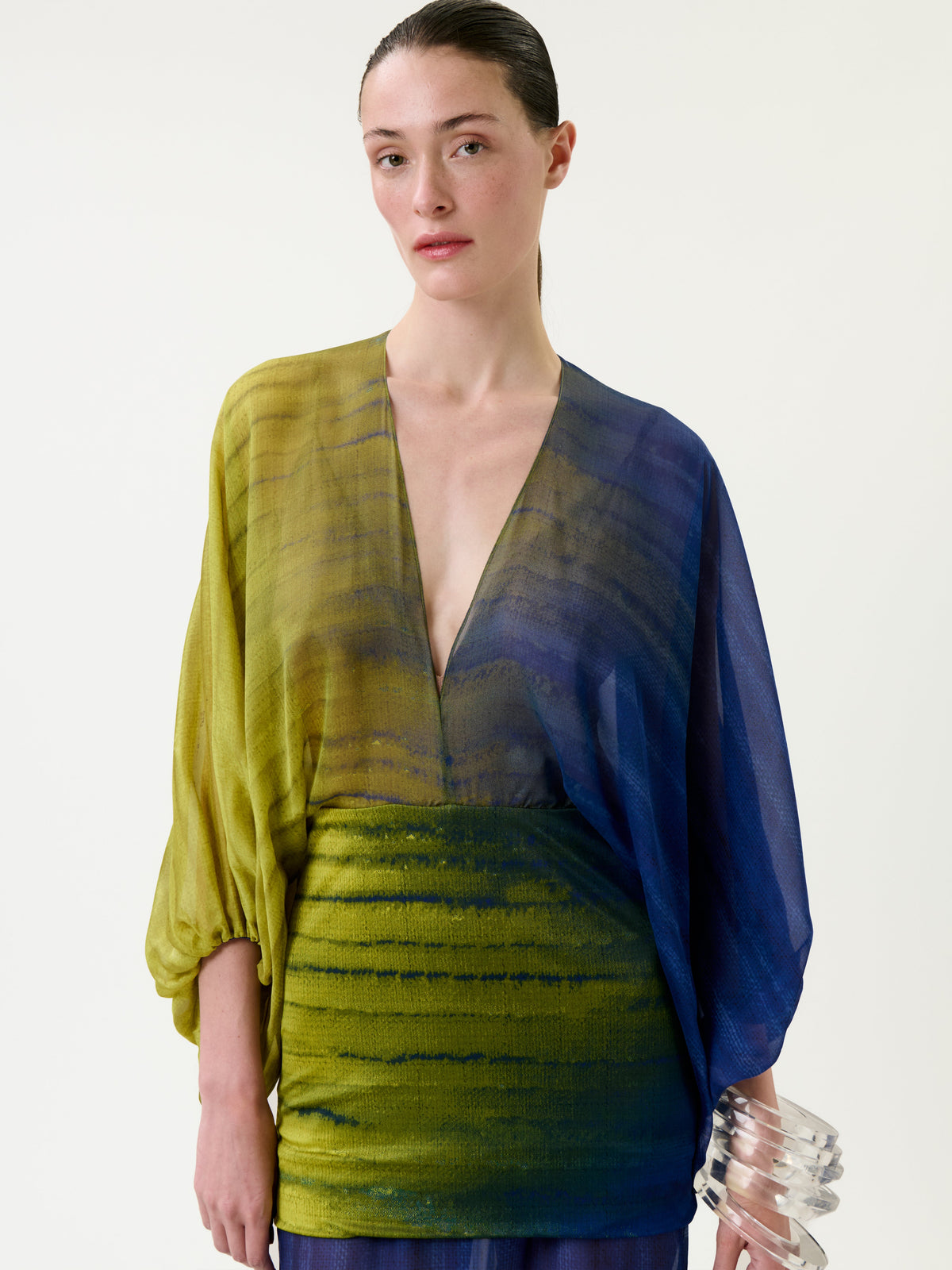 A woman wearing the Lilen Dress—a sheer design with a deep V-neck and green-to-blue gradient tones—stands against a plain background, holding a translucent, wavy bracelet in her right hand.