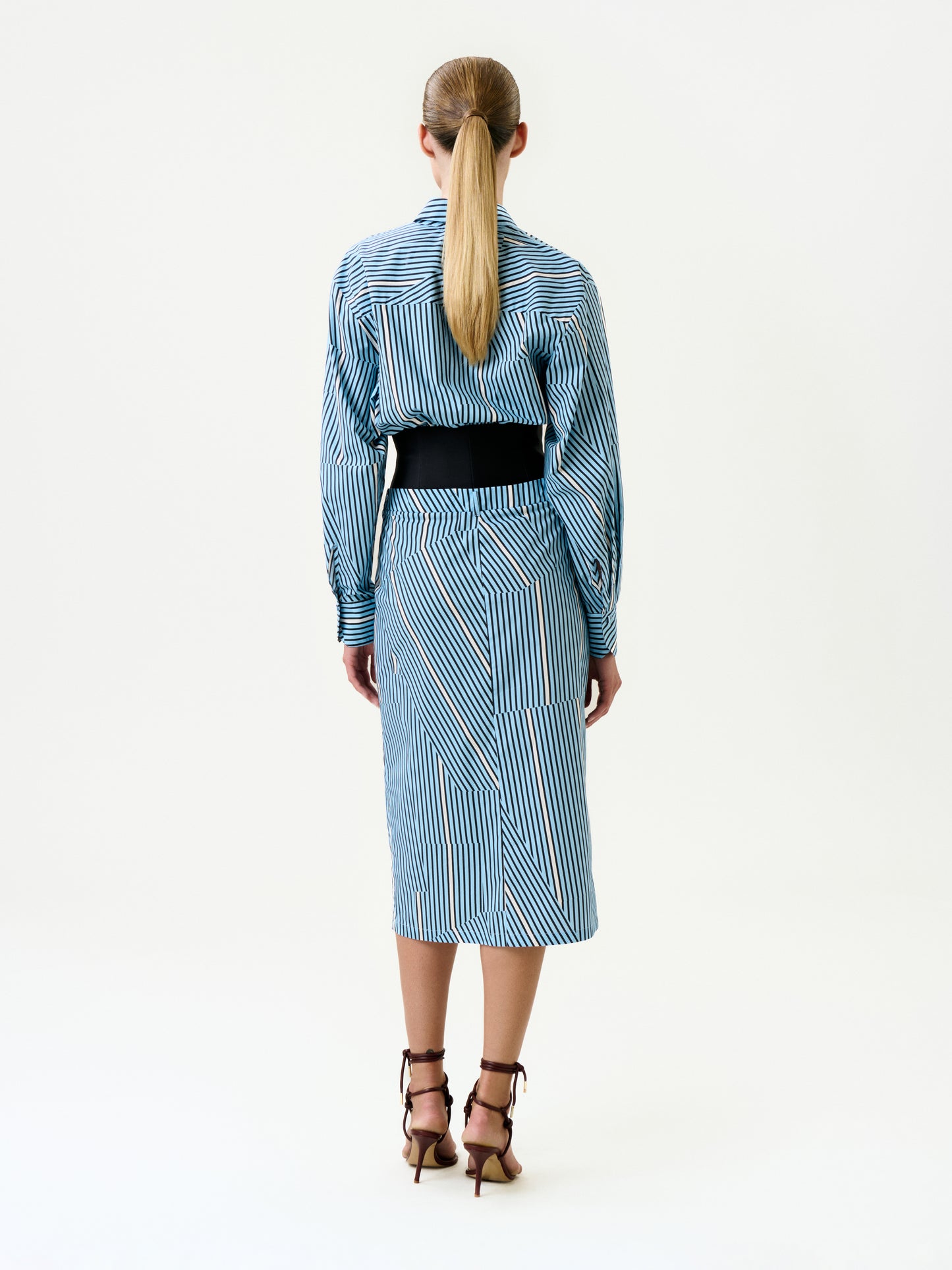A person with long blonde hair in a low ponytail stands with their back to the camera, wearing the Lorena Dress—a blue and white striped 100% cotton midi dress—cinched with a black belt and paired with brown high-heeled sandals.