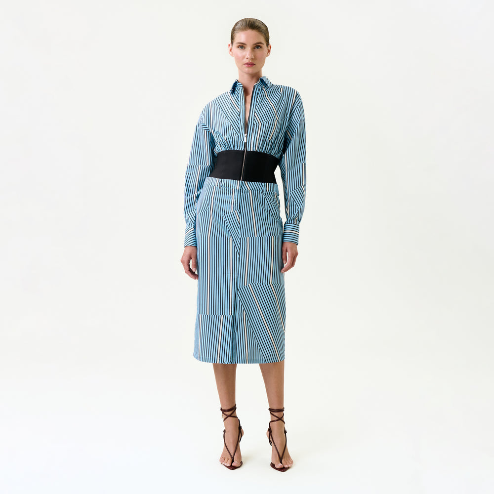 A woman poses against a white background in the Lorena Dress, a blue and white vertical striped midi made from 100% Italian cotton, with long sleeves, a black wide belt, and strappy brown high-heeled sandals.