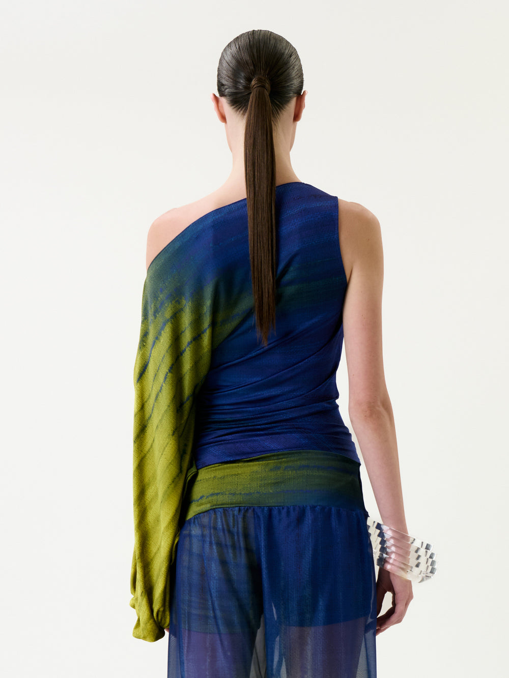 A person with long dark hair in a ponytail stands with their back to the camera, wearing the Rhona Blouse—a blue and green one-sleeve top—paired with a sheer blue skirt. On their right wrist, they wear a chunky, translucent bracelet.