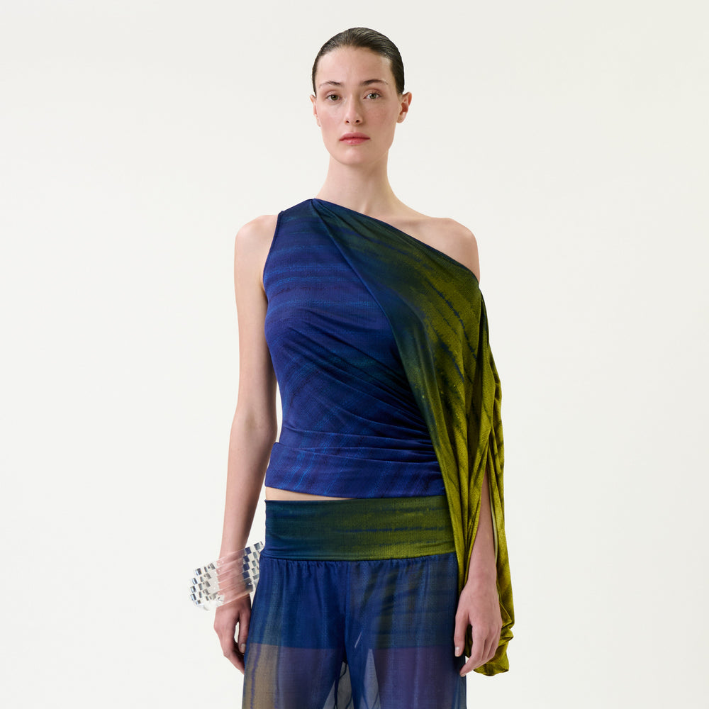 A person stands against a plain background, wearing the sheer, asymmetrical Rhona Blouse in blue and green with one shoulder exposed, paired with matching sheer pants. Their hair is slicked back, and they hold a small, beaded accessory.