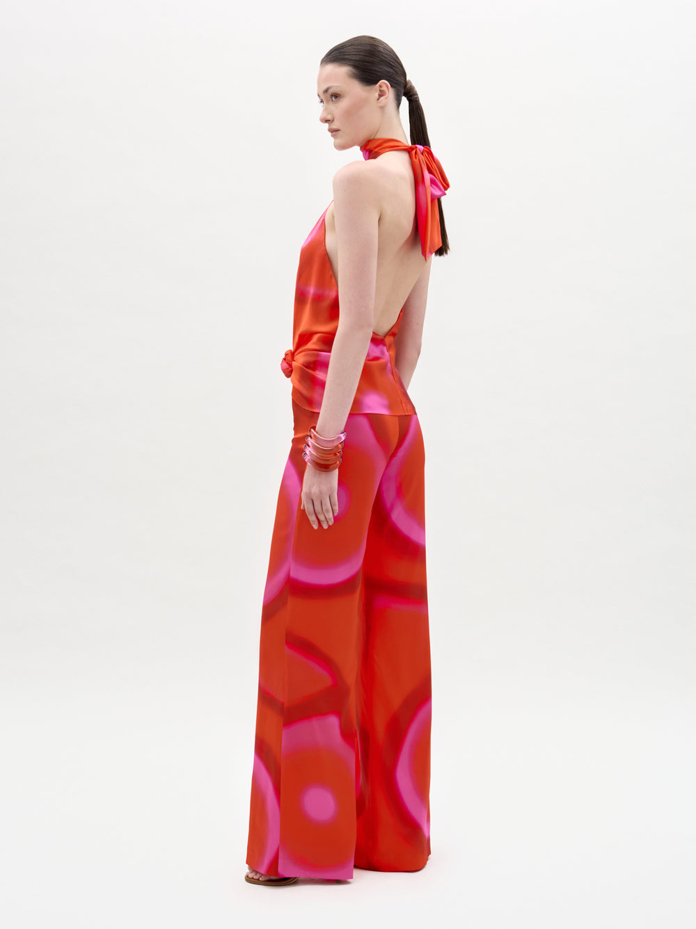 A woman stands sideways against a white background, looking over her shoulder while wearing the Andie Pant—a red and pink halter-neck backless jumpsuit with high-waisted wide-leg pants. Her straight brown hair is tied back and she accessorizes with red bangles.