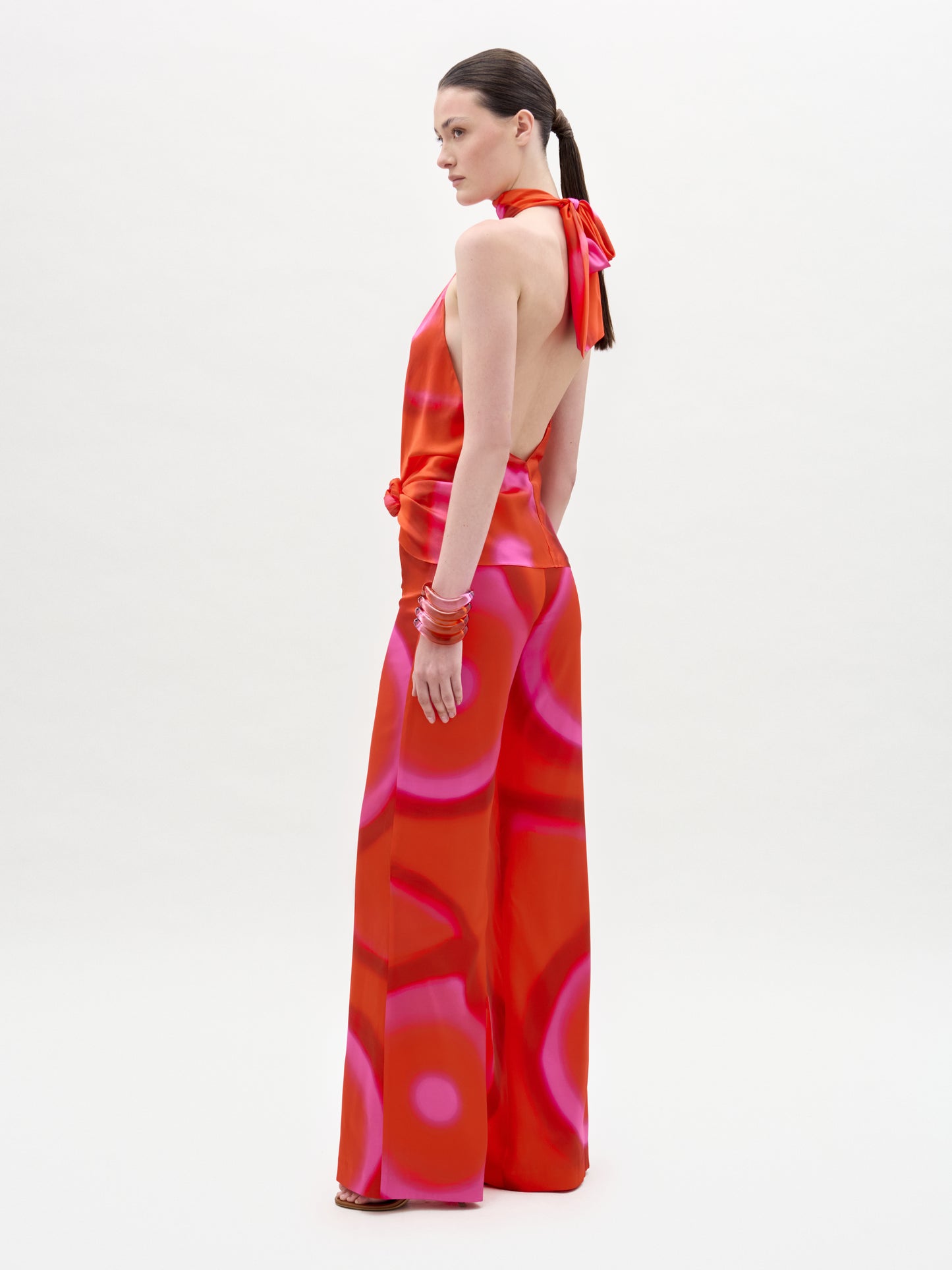 A woman stands sideways against a white background, looking over her shoulder while wearing the Andie Pant—a red and pink halter-neck backless jumpsuit with high-waisted wide-leg pants. Her straight brown hair is tied back and she accessorizes with red bangles.
