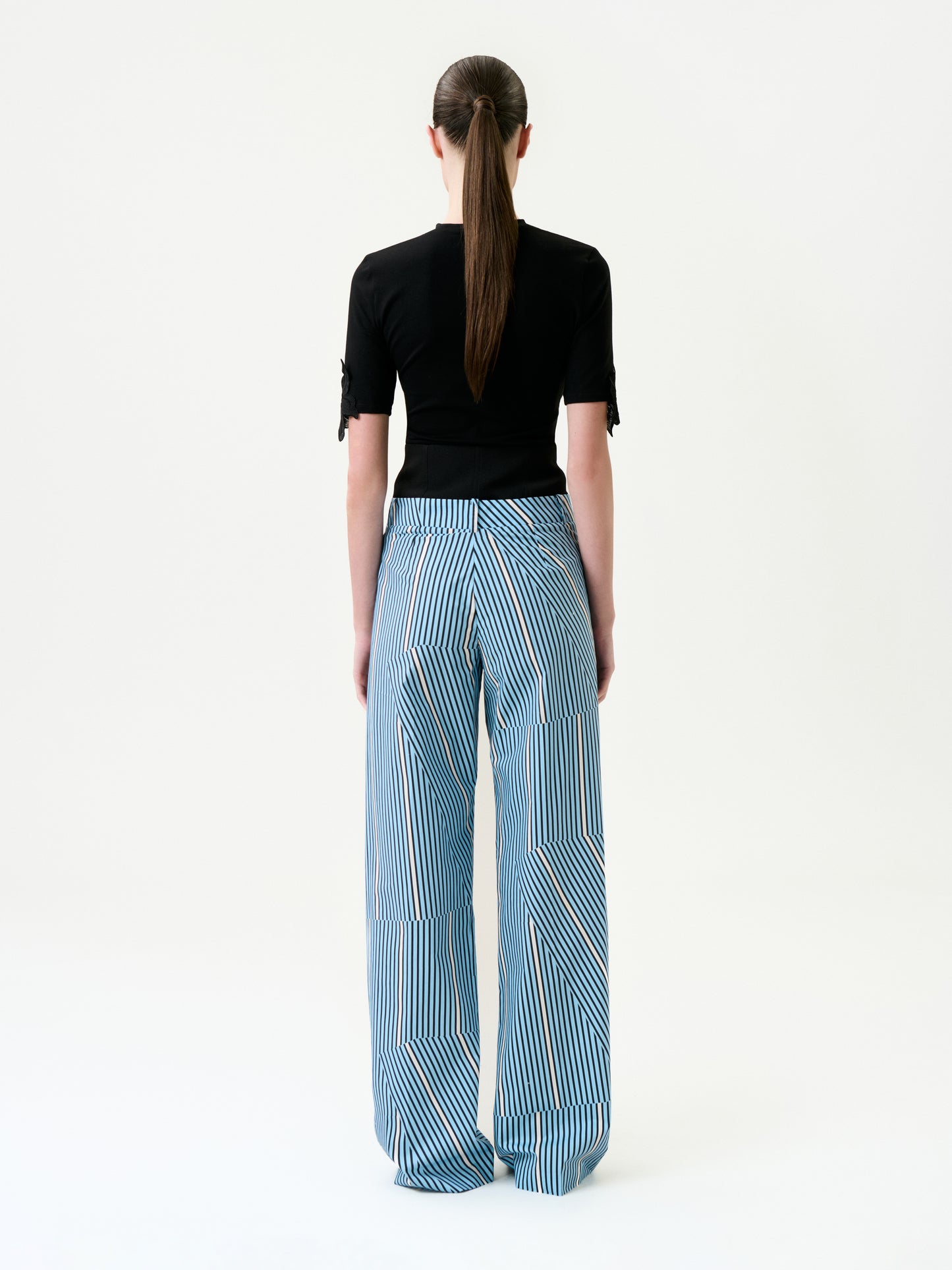 A person with long brown hair in a low ponytail faces away, wearing a fitted black short-sleeve top and Ruelle Pant—wide-leg blue pants with vertical and diagonal white stripes—crafted from luxurious Italian fabric, against a white background.