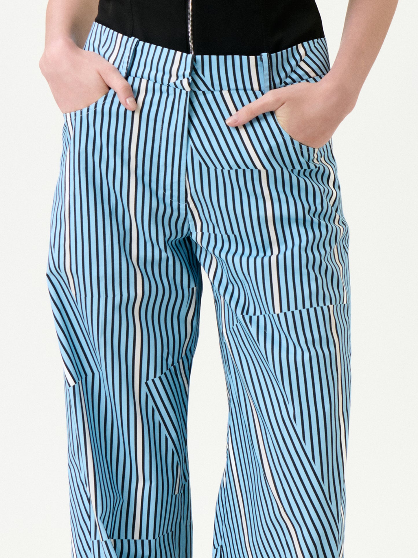 Someone wears the Ruelle Pant—a pair of blue and white vertically striped pants made from 100% cotton—with hands in pockets, shown from waist to mid-thigh against a plain background.