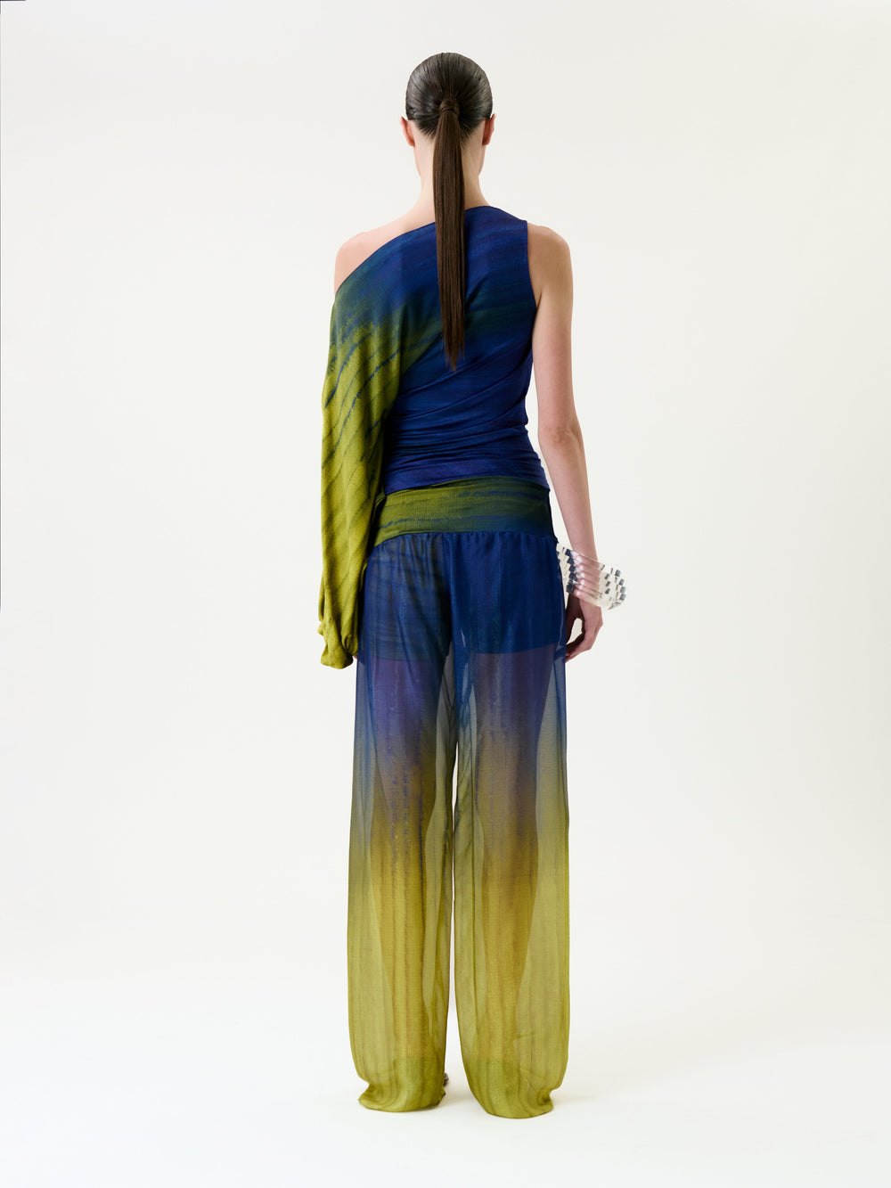 A person with long hair in a ponytail models the Lene Pant—a sheer, blue and yellow ombre pant—paired with a matching one-sleeve top, standing against a plain white background and facing away from the camera.