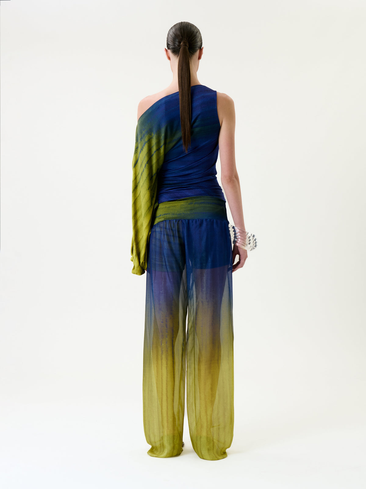 A person with long hair in a ponytail models the Lene Pant—a sheer, blue and yellow ombre pant—paired with a matching one-sleeve top, standing against a plain white background and facing away from the camera.
